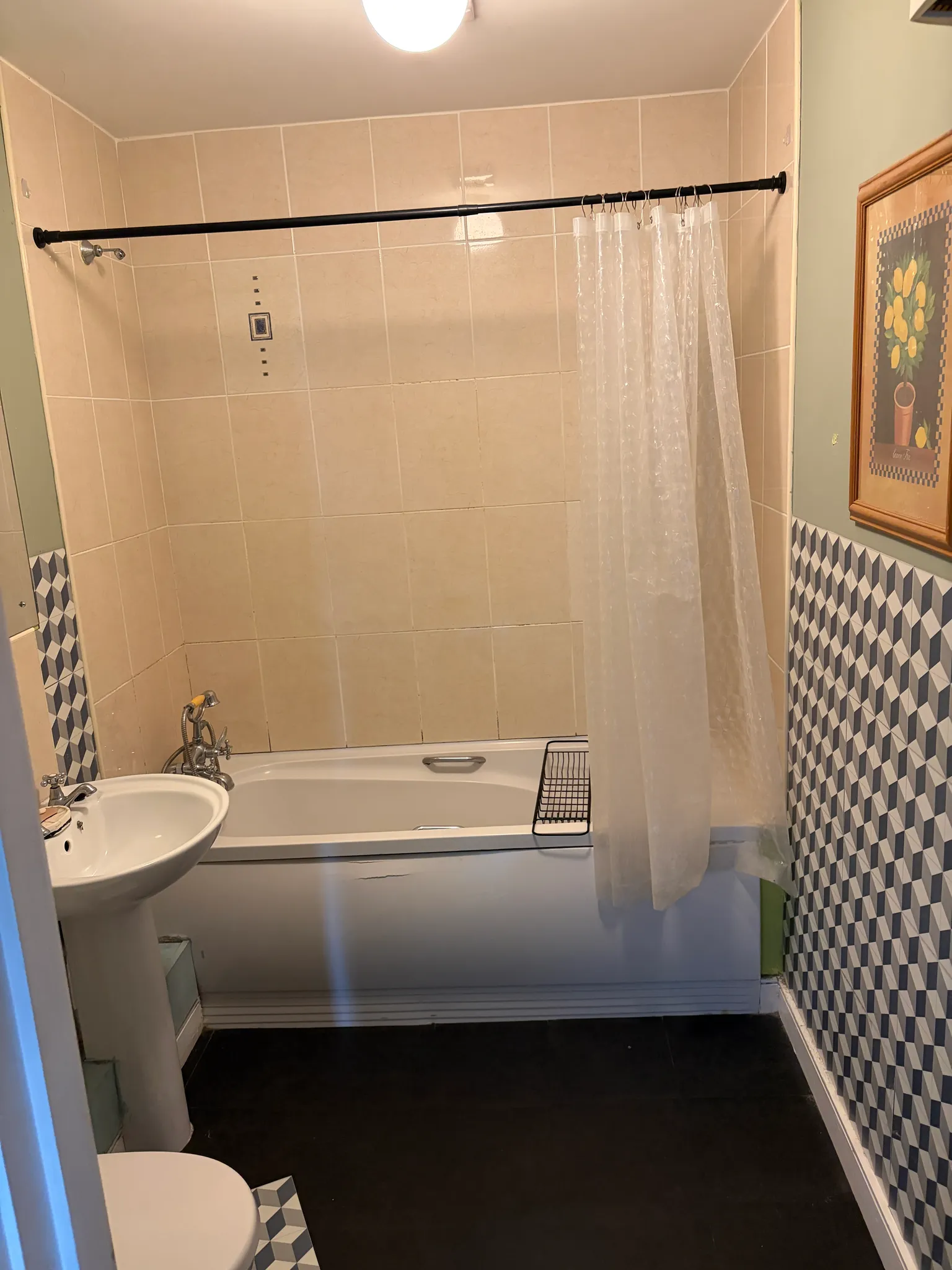 Bathroom with a bathtub, clear shower curtain on a rod, wall tiles, pedestal sink and partial view of a toilet and framed artwork.