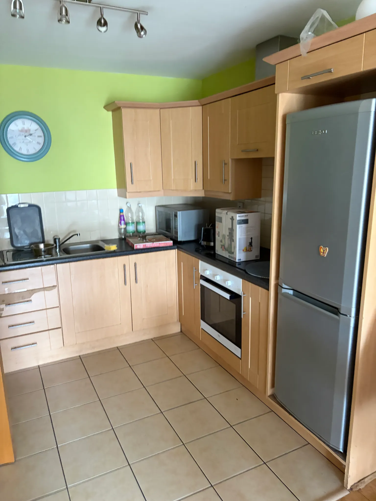 Kitchen with light wood cabinets, tiled floor, stainless refrigerator, oven, microwave and sink with a green wall and a round wall clock.