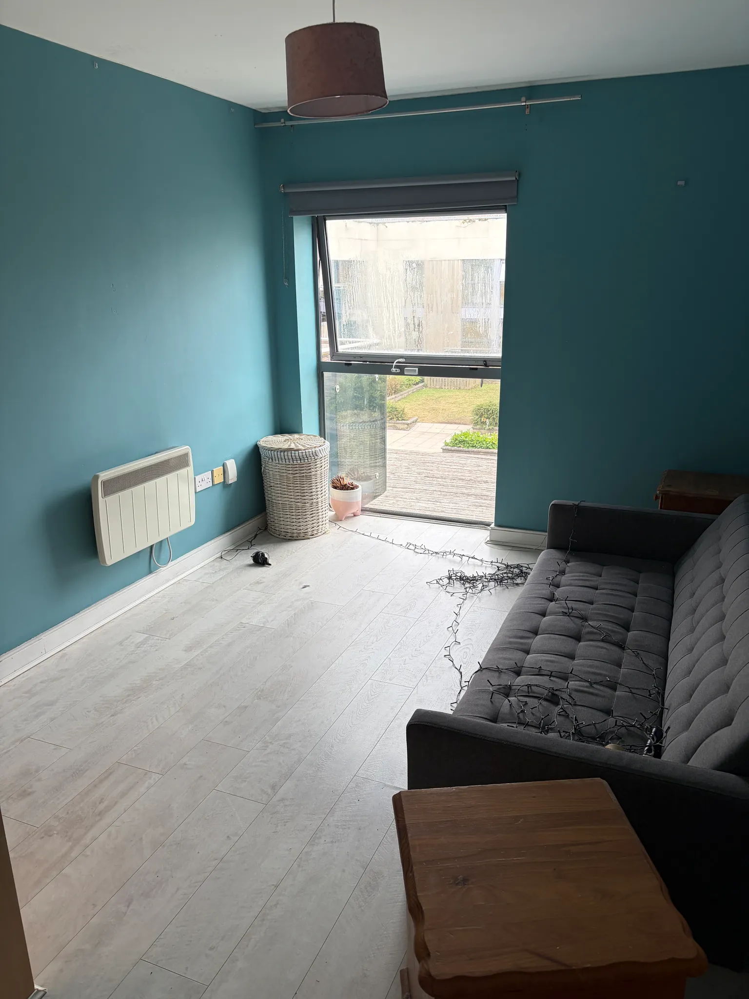 Living room with a grey sofa, large window with a roller blind, blue painted walls, light wood floor and a small wooden side table.
