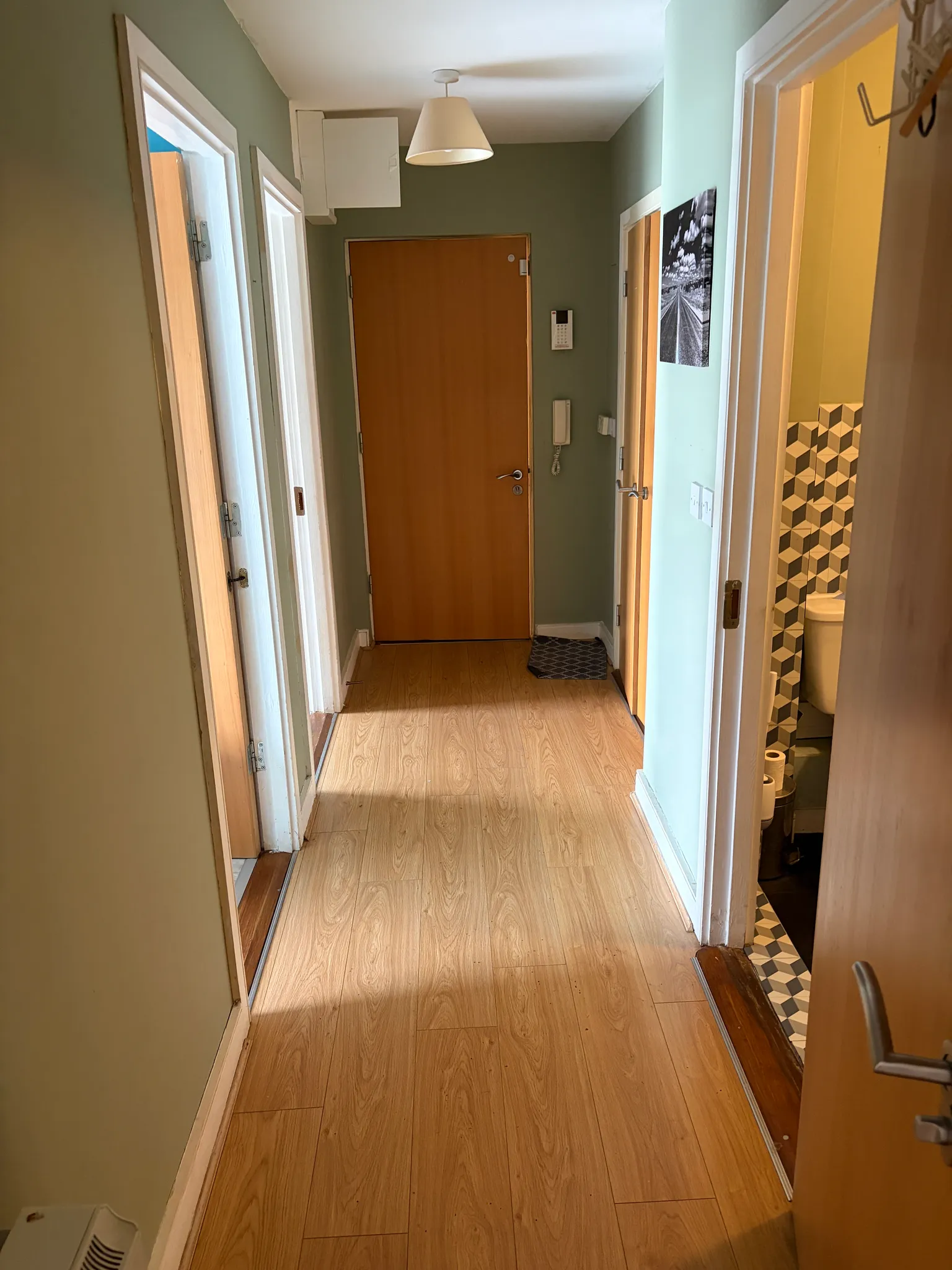 Hallway with a closed wooden entrance door, light wood floor, open doorways to adjacent rooms and a visible bathroom with patterned tiles.