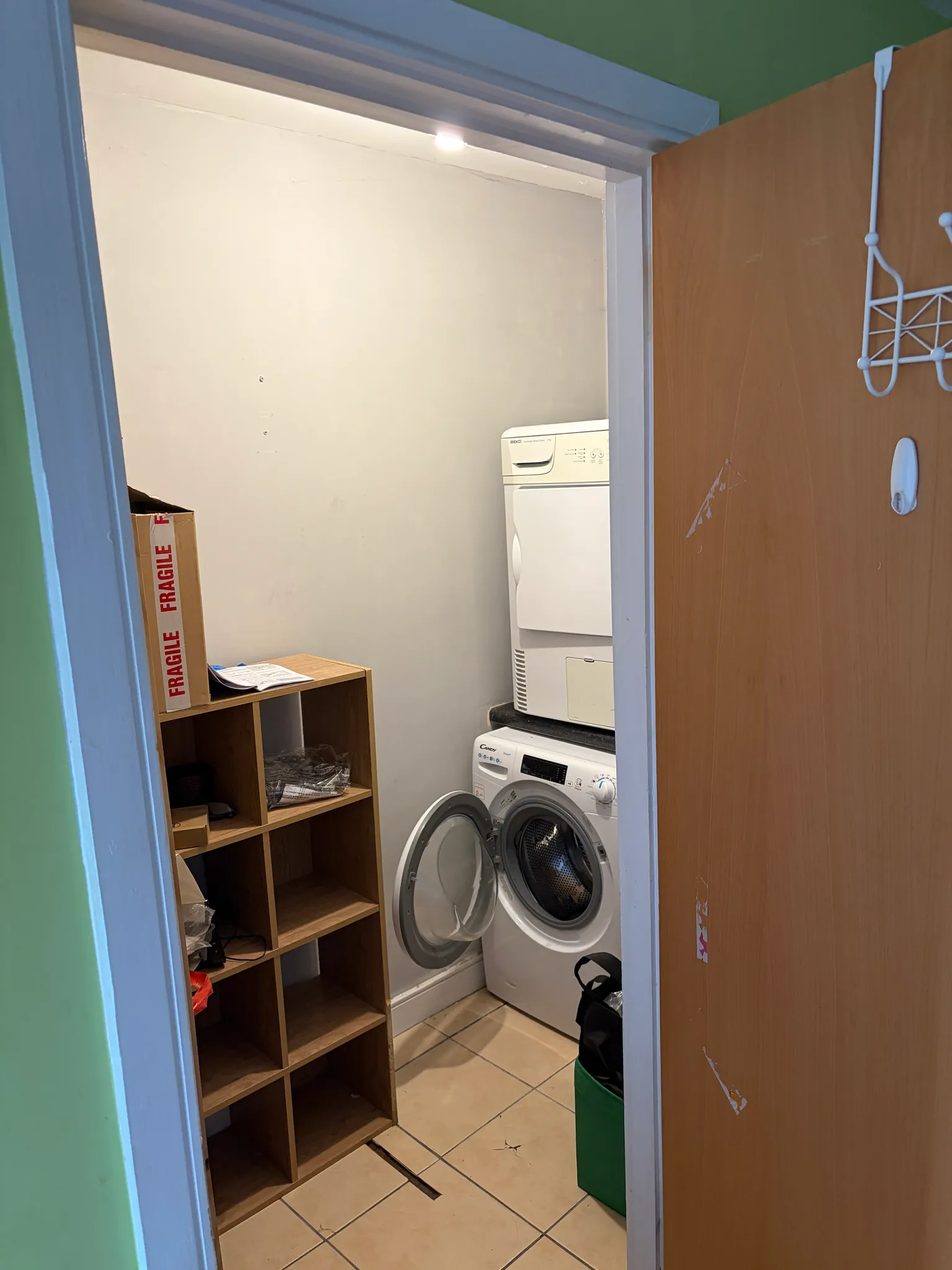 Small laundry room with a stacked dryer and front-load washing machine (washer door open), wooden cubby shelf and tiled floor, viewed through a partly open door.