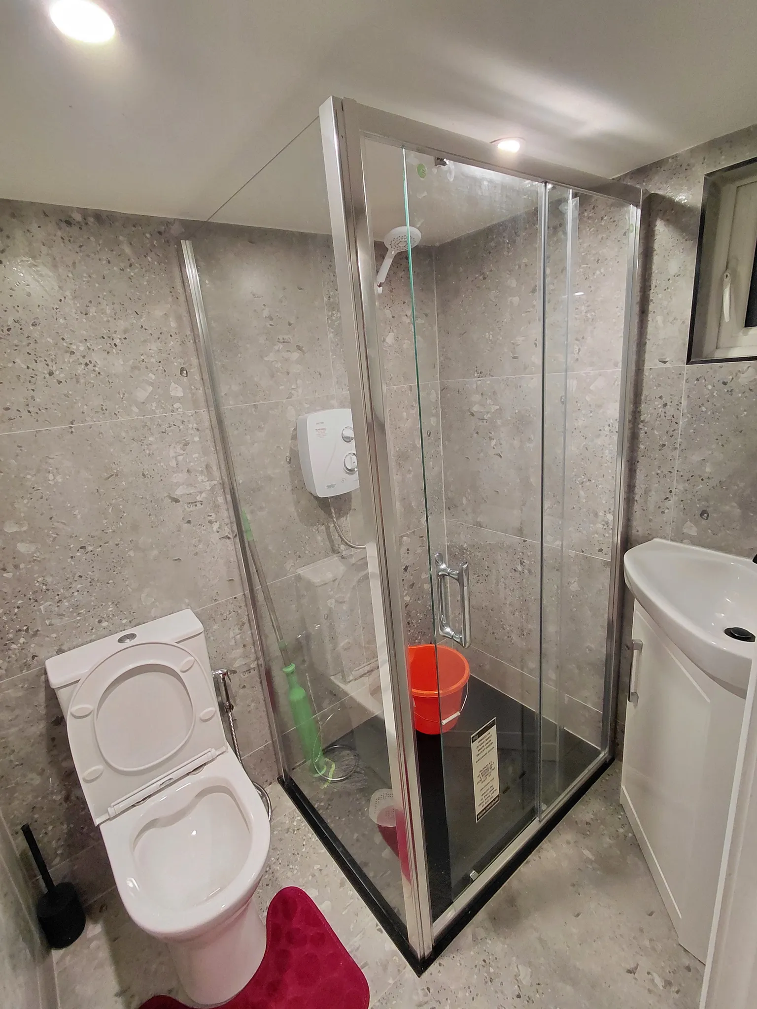Bathroom with a glass-enclosed corner shower, wall-mounted shower unit, toilet and small sink.