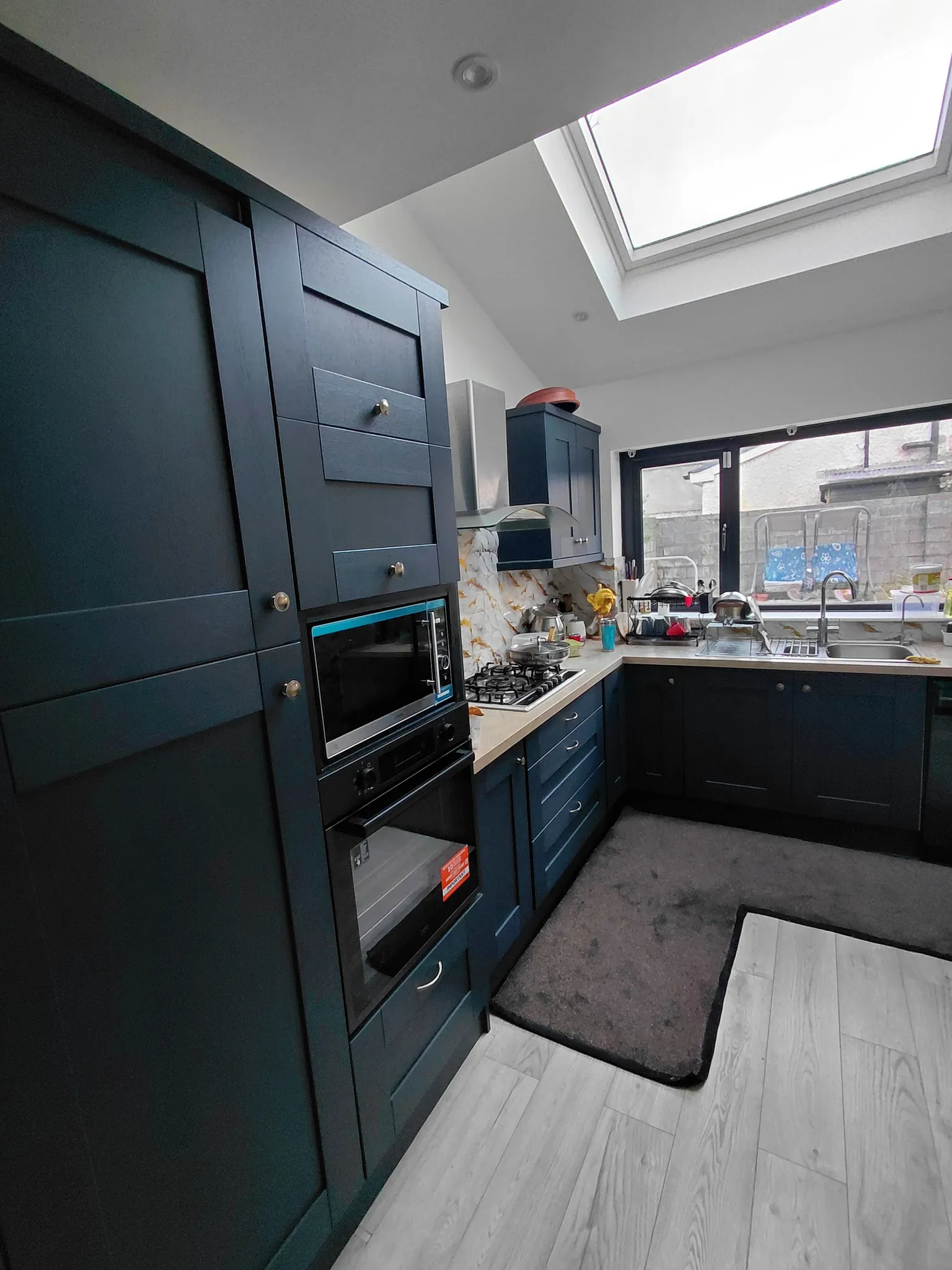 Kitchen with dark blue cabinets, built-in oven and microwave, gas hob with hood, countertop sink and a large skylight window above.