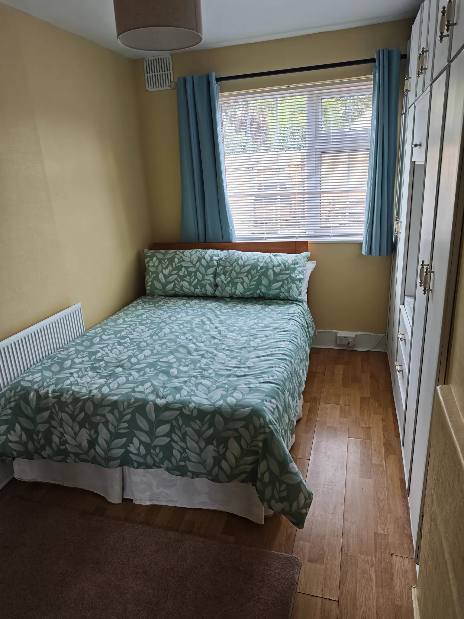 Bedroom with a double bed with patterned duvet, window with horizontal blinds and blue curtains, built-in wardrobe and radiator.
