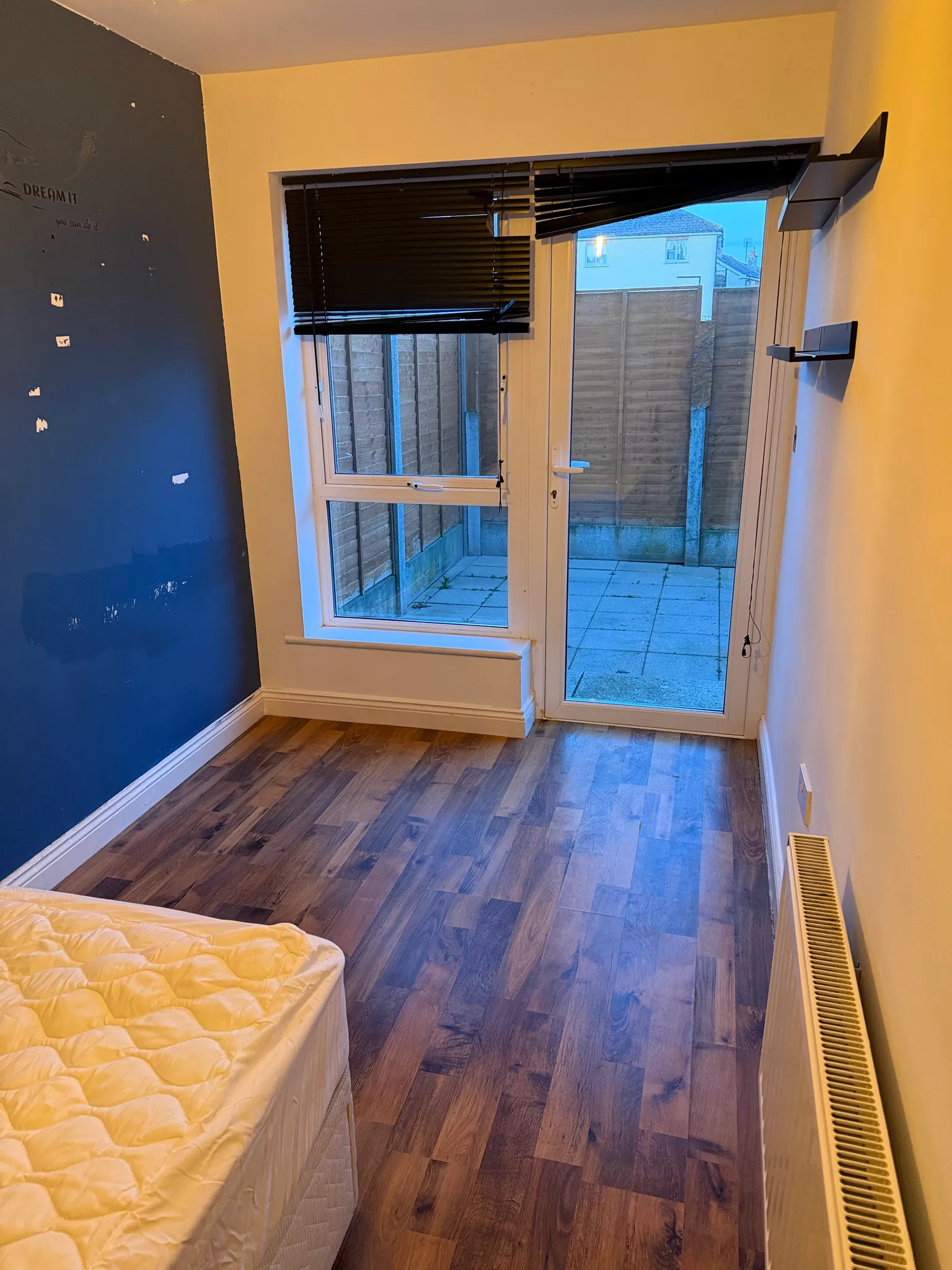 Bedroom with a mattress at the lower left, wooden floor, a window and glass door with partially closed blinds leading to a small paved outdoor area, radiator on the right and wall-mounted shelves on the right wall.