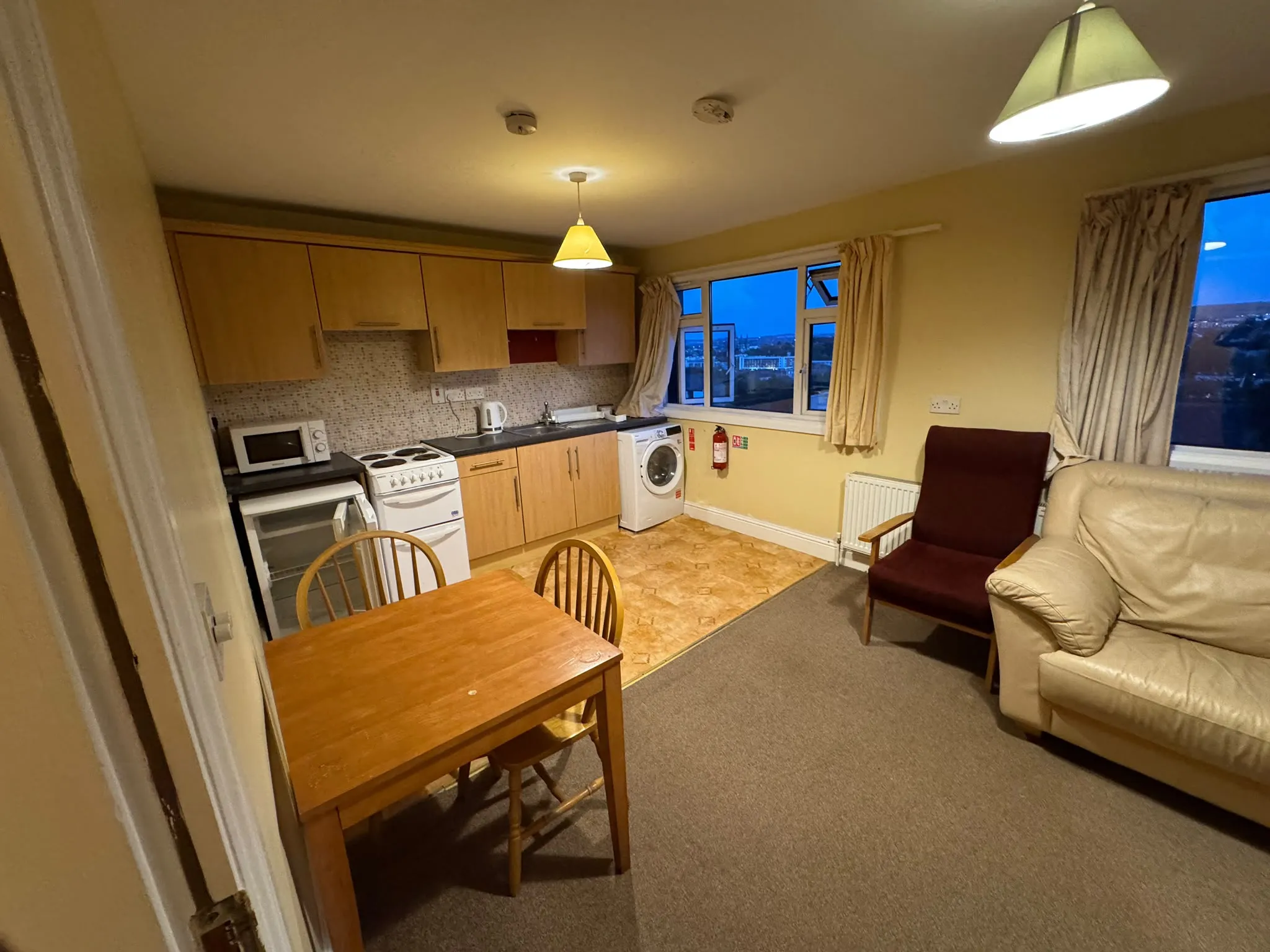 Open-plan living area with a small kitchenette, dining table and chairs, washing machine under the counter, stove and microwave, two armchairs and windows with curtains.