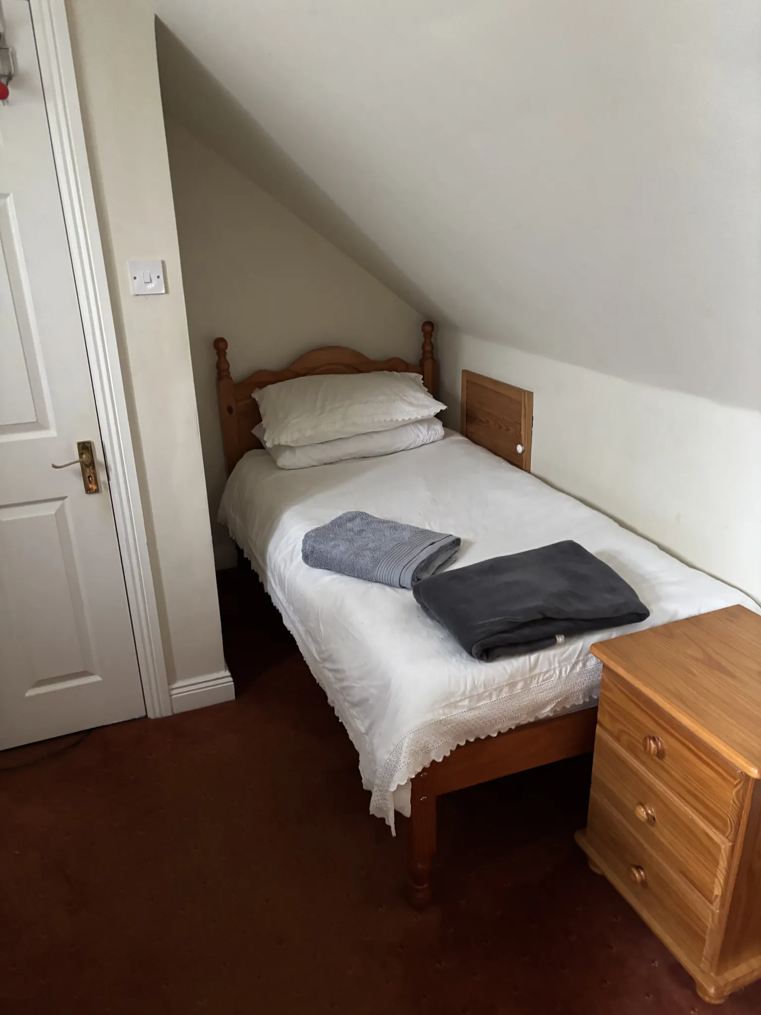 Small bedroom with a single bed positioned under a sloped ceiling, white bedding and pillows, folded towels on the bed, and a wooden bedside chest and door visible.
