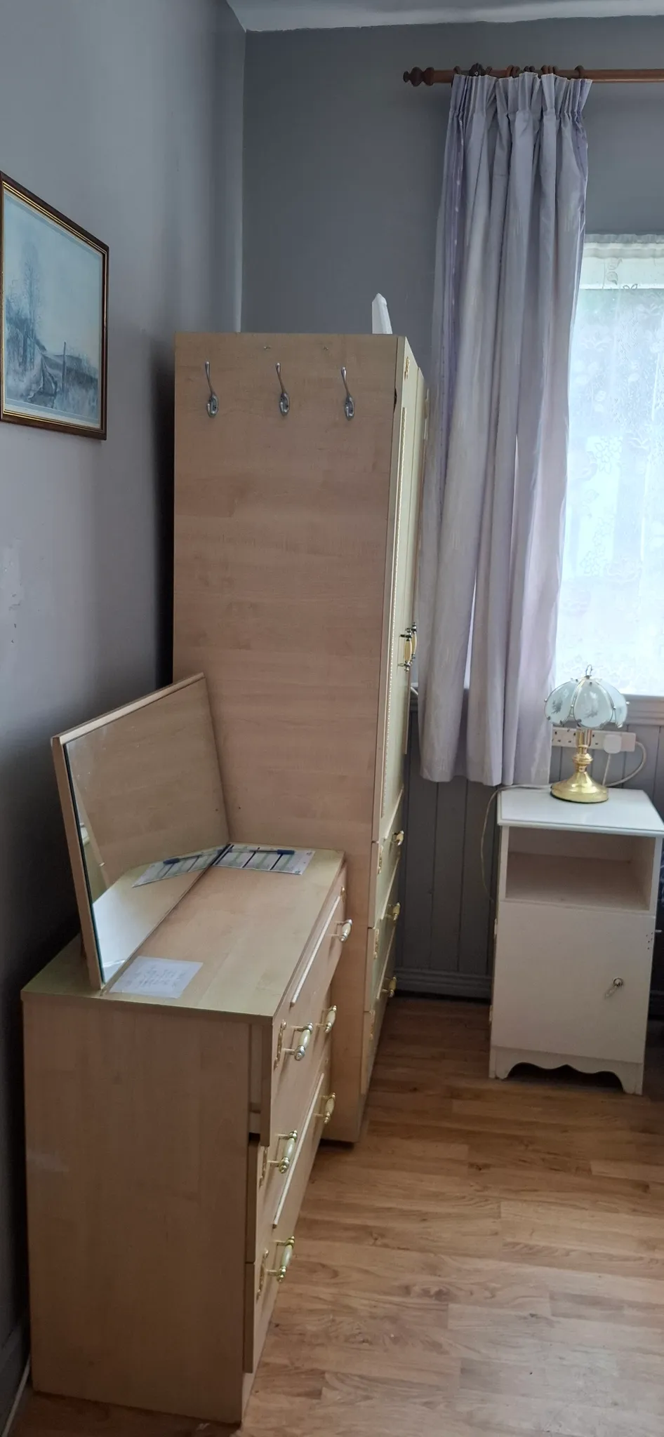 Bedroom corner with a wooden dresser and matching tall wardrobe, a small white bedside cabinet with lamp, a mirror on the dresser, a window with curtains, and a framed picture on the wall.