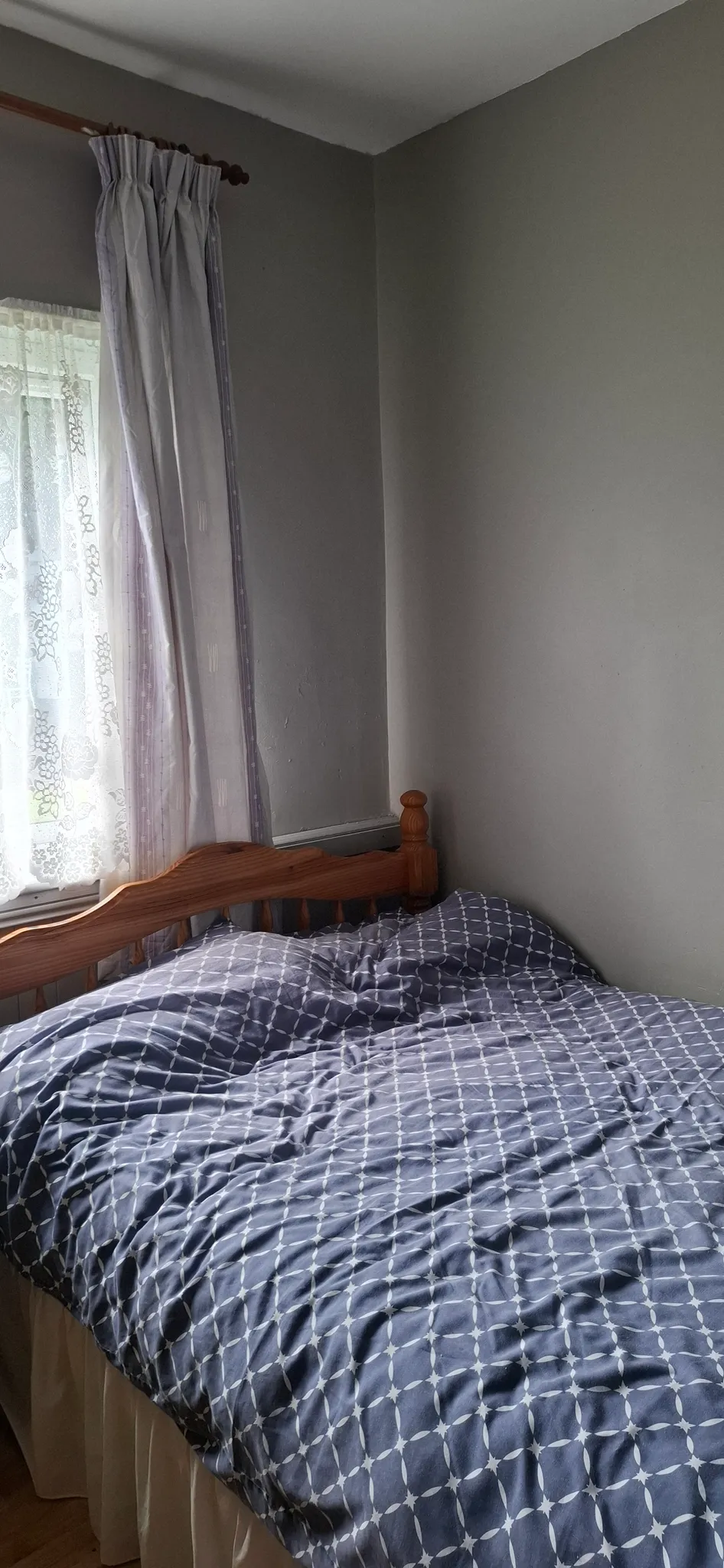 Bedroom corner with a wooden single bed, patterned duvet, and a window with lace and fabric curtains.