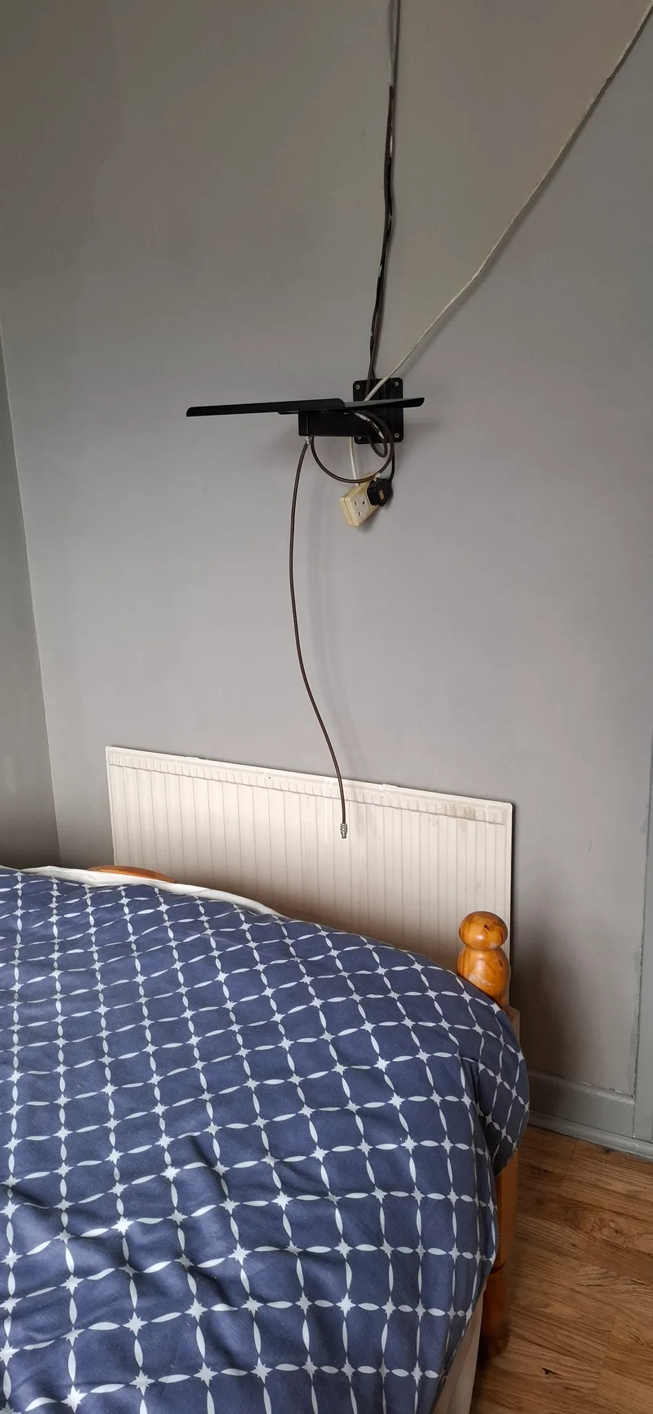 Bedroom corner showing a bed with patterned duvet, radiator on the wall, and a wall-mounted TV bracket with hanging cables and plug.