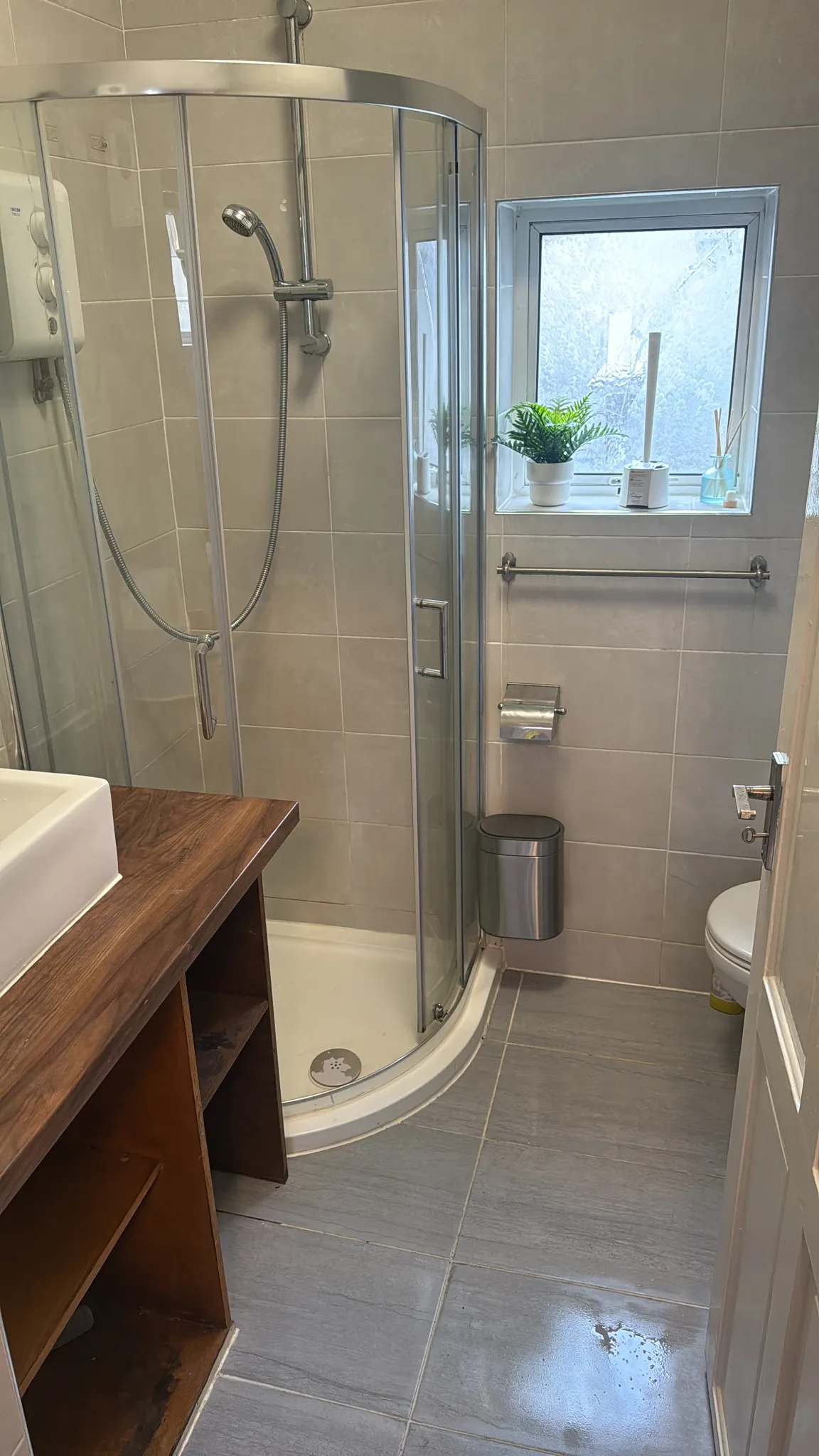 Bathroom with a corner glass shower enclosure, handheld showerhead, small window with potted plant, sink countertop and toilet.