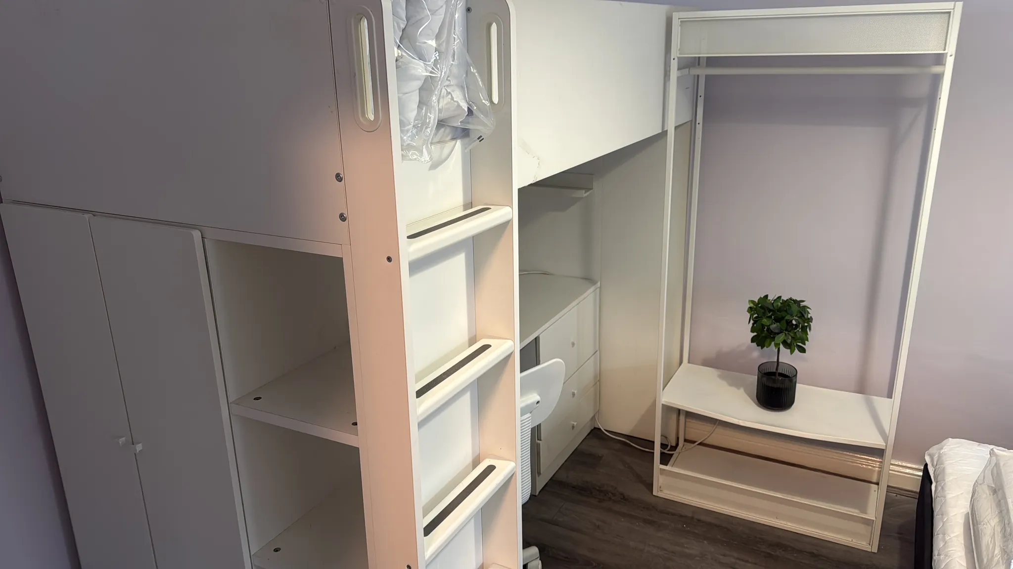 Bedroom corner showing a lofted bed frame with ladder, built-in shelving and a small open wardrobe with a desk and a potted plant.