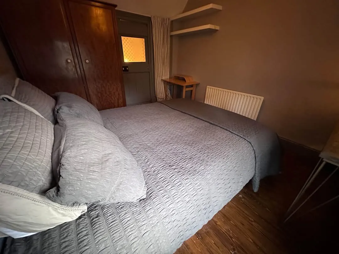 Bedroom with a double bed covered by a gray quilt and pillows, a wooden wardrobe, radiator, small desk, shelves and a door with a curtained window.