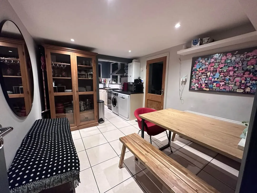 Dining area with a wooden table, bench and red chair, open view into a small kitchen with washing machine and cabinets, wall mirror and framed artwork on tiled floor.