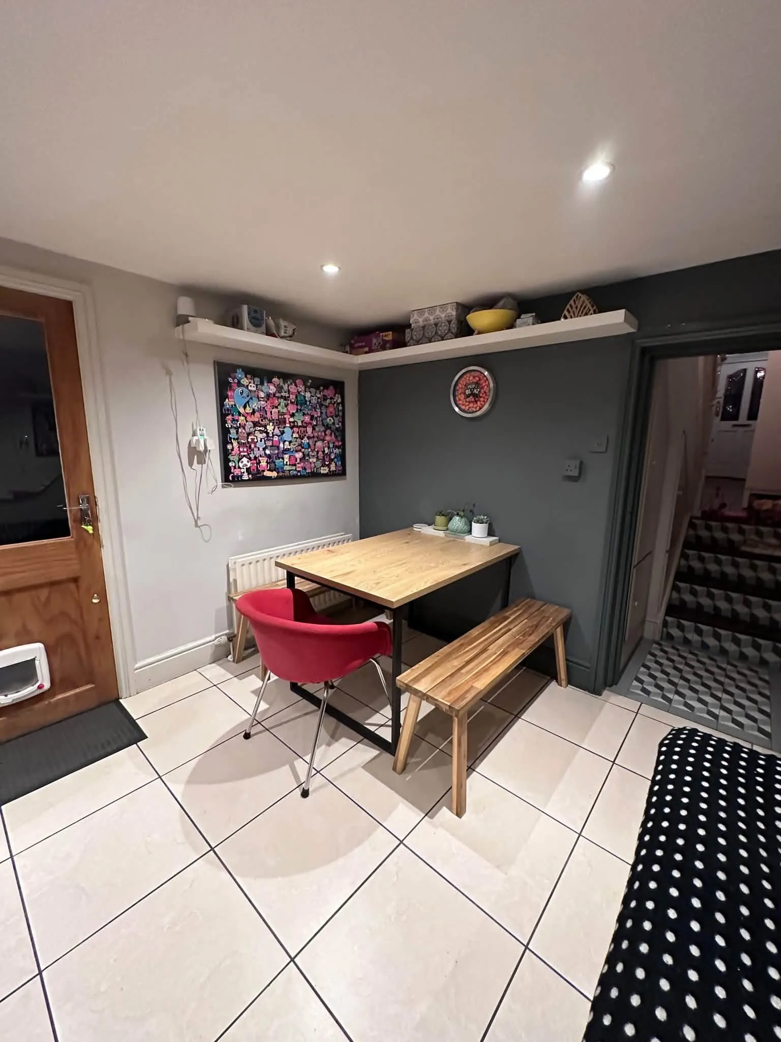 Dining area with a wooden table, red upholstered chair, wooden bench, tiled floor, wall-mounted shelf and framed artwork.