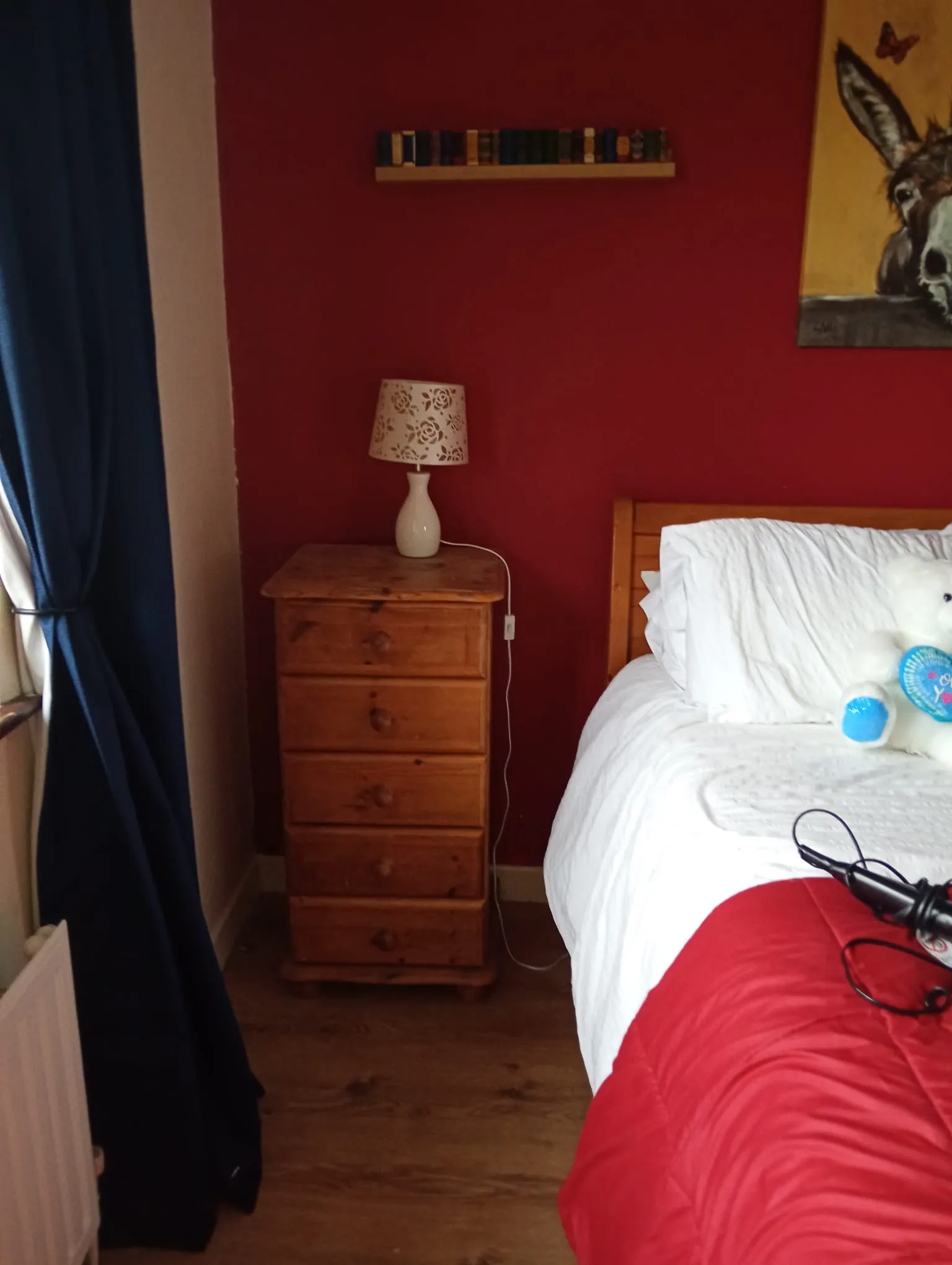 Bedroom corner with a wooden bedside chest, table lamp, and part of a bed with white bedding against a red wall