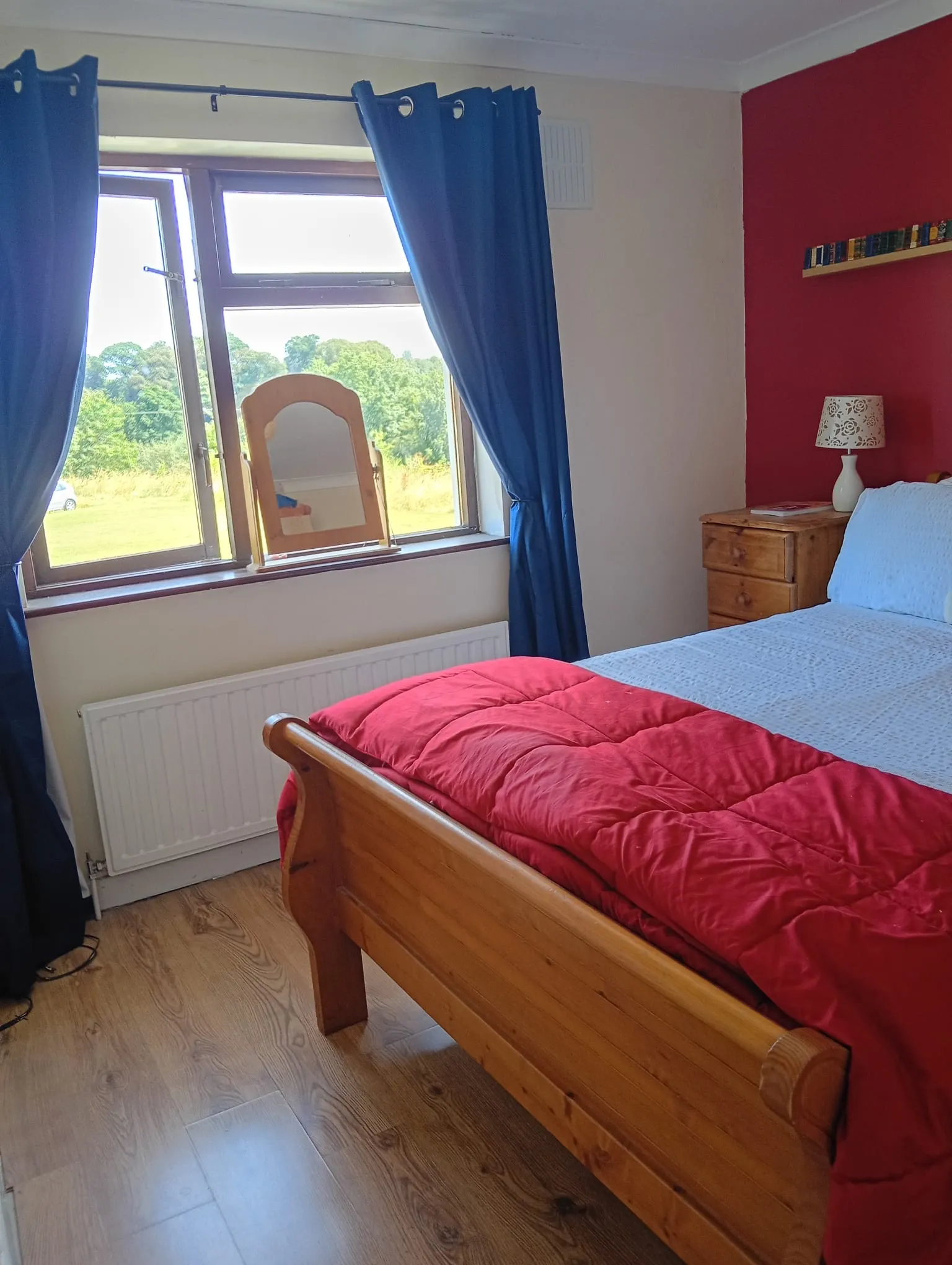 Bedroom with a wooden bed, red duvet, window with blue curtains and a small wooden bedside table with lamp