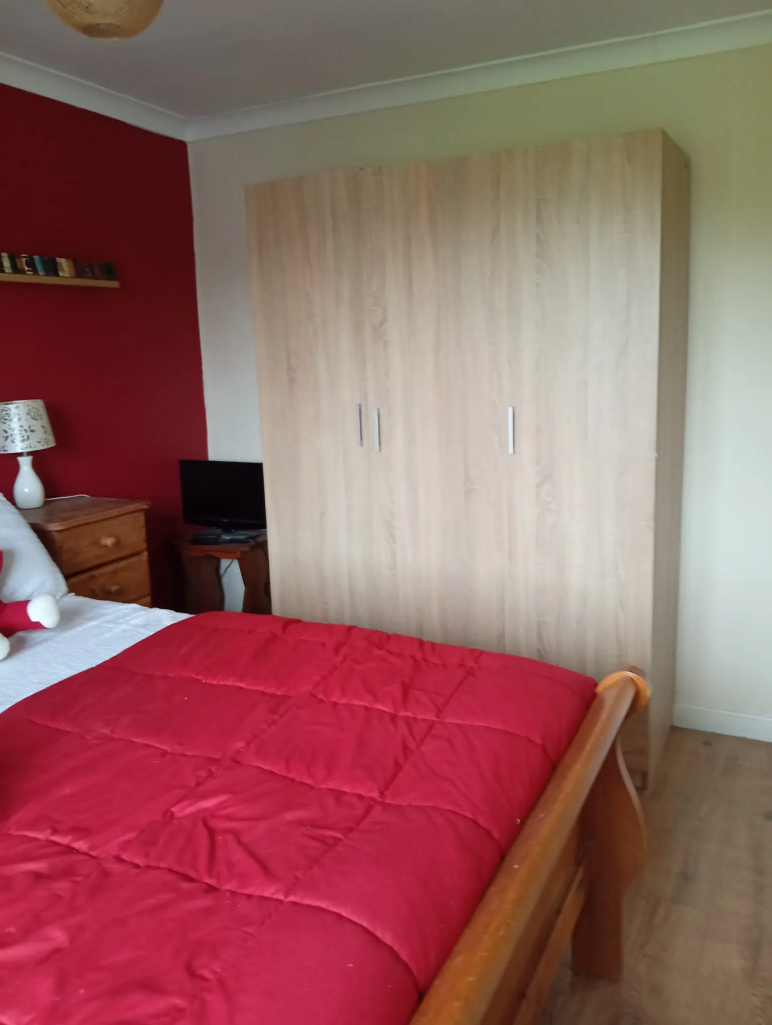 Bedroom with a red duvet on a wooden bed, a large light-wood wardrobe, a bedside table with a lamp and a small TV, and a red accent wall