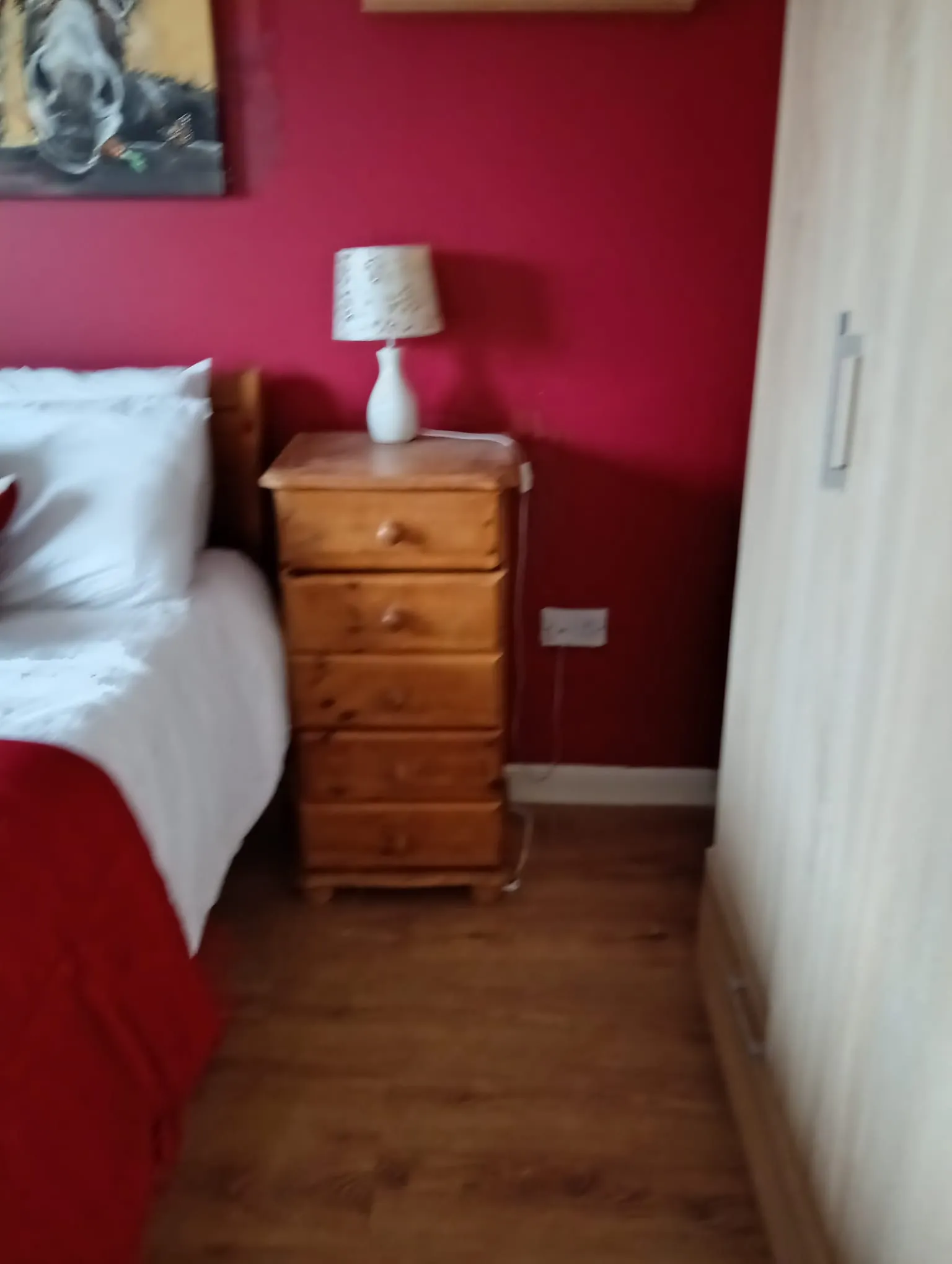 Bedroom corner showing a bed edge with white bedding, a wooden bedside chest with a table lamp, a red painted wall and a wardrobe at the right.
