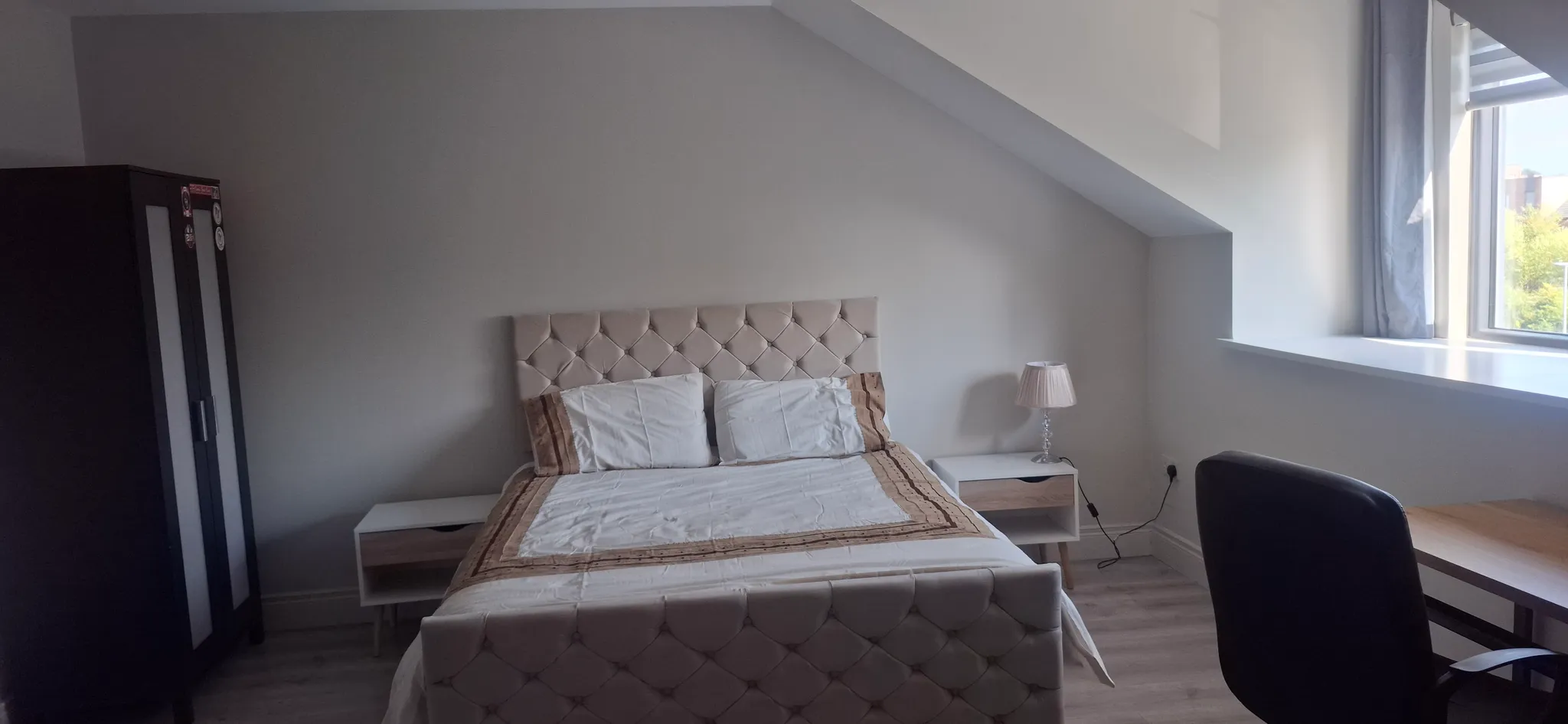 Bedroom with a double bed and upholstered headboard centered against a sloped wall, two bedside tables (one with a lamp), a wardrobe to the left, and a desk and chair by a window to the right.