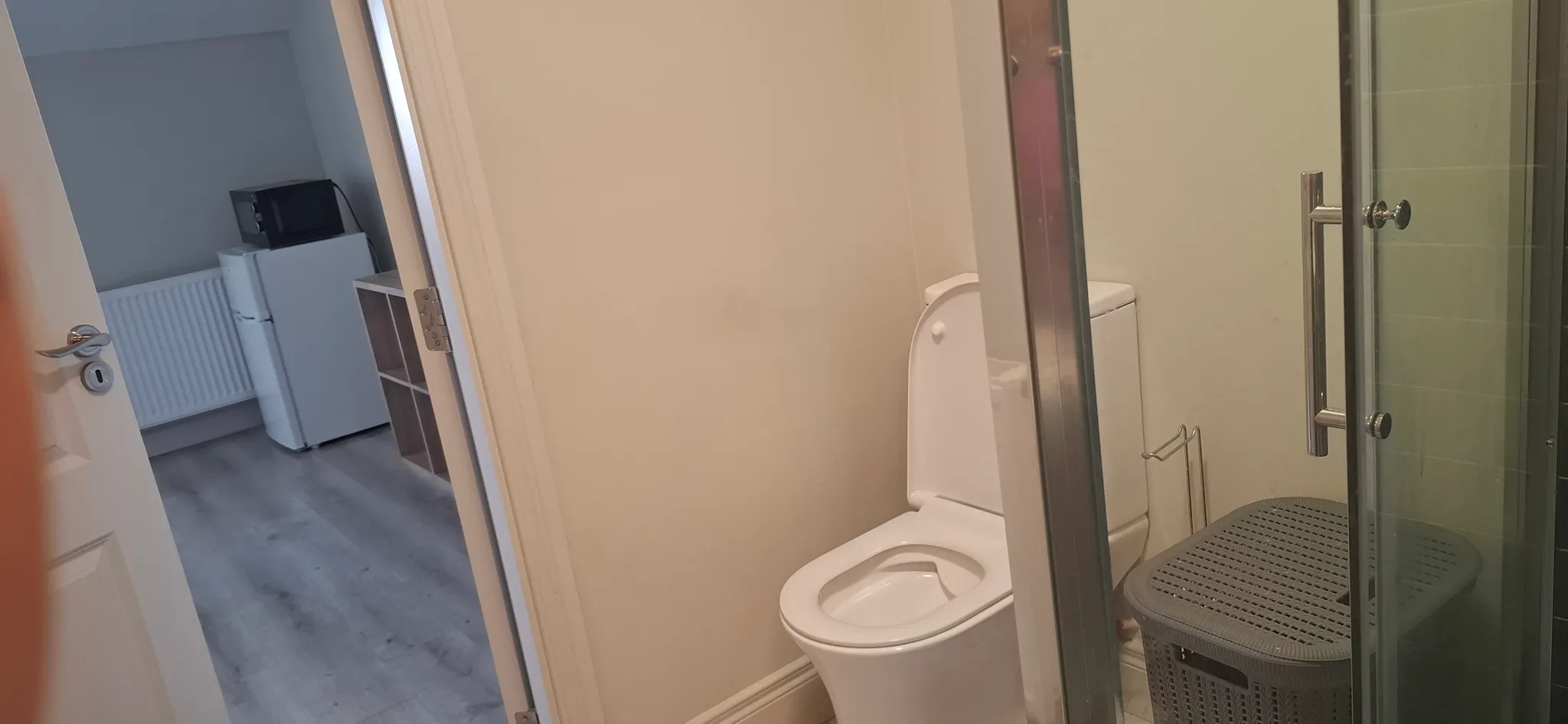 Bathroom with a toilet, glass shower door, and a plastic laundry basket; view through the open door into an adjacent room showing a small refrigerator and microwave.