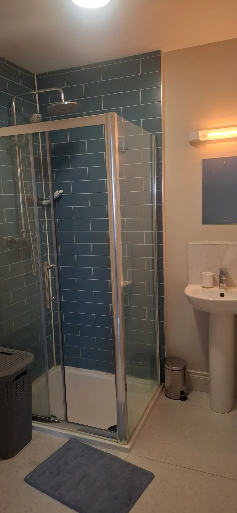 Bathroom with a corner glass shower enclosure, blue subway tile wall, overhead showerhead, pedestal sink and small trash bin.