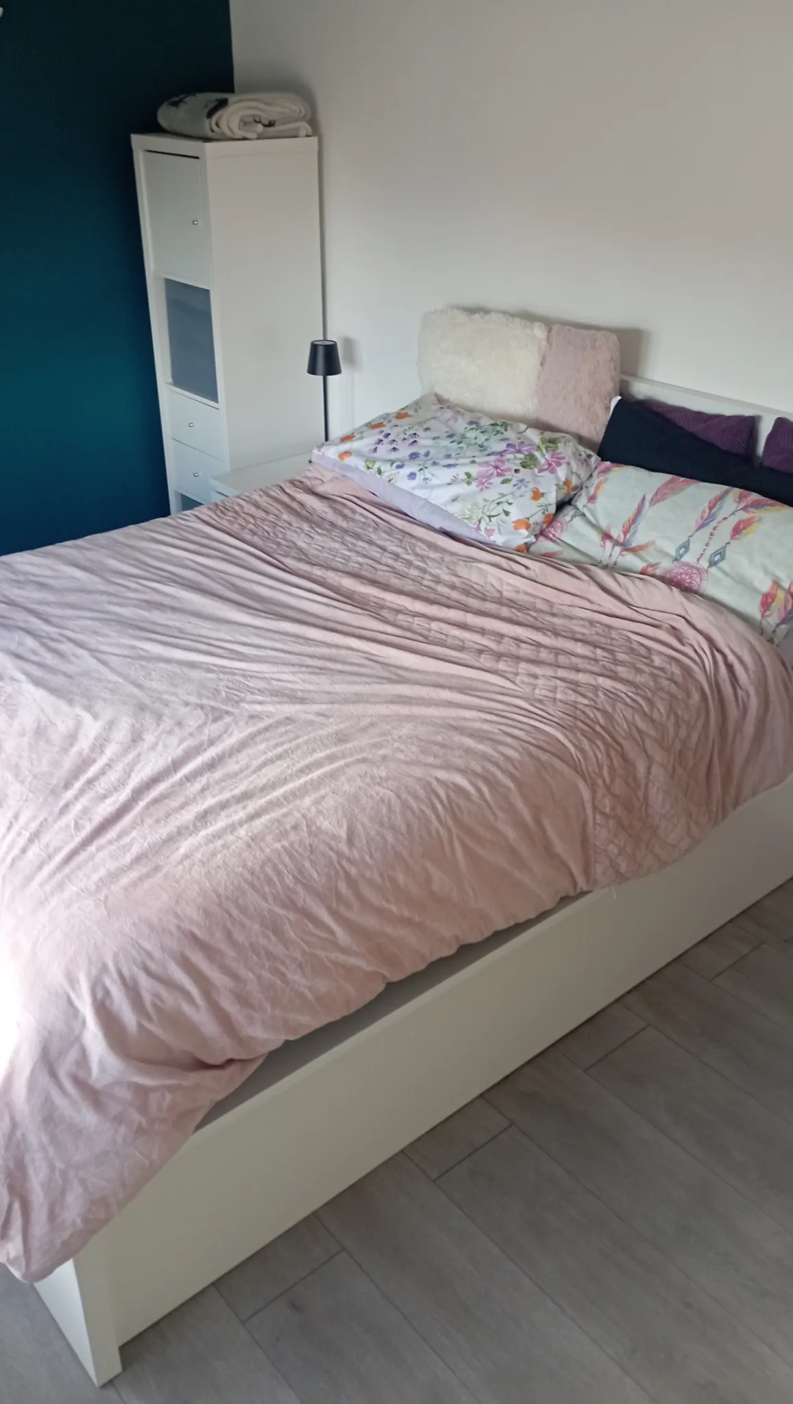 Bedroom with a double bed covered by a pink duvet, several pillows, a small black bedside lamp and a white shelving unit against a blue and white wall.