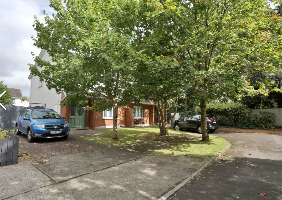 Exterior view of a house with a driveway, two parked cars, trees, a small lawn, and a front door.