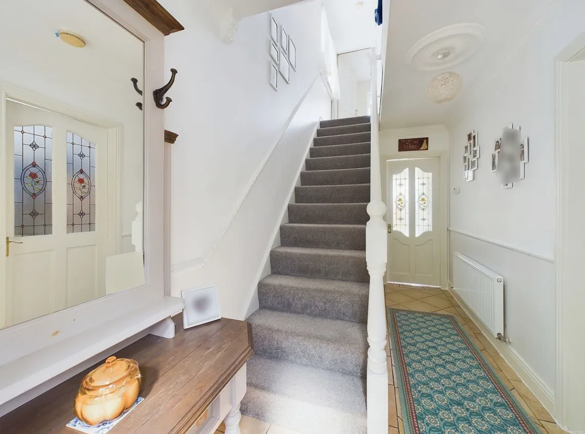 Hallway with a carpeted staircase up the center, wooden console table with a decorative jar and mirror on the left, patterned runner rug, radiator on the right, and a front door with stained-glass panels.