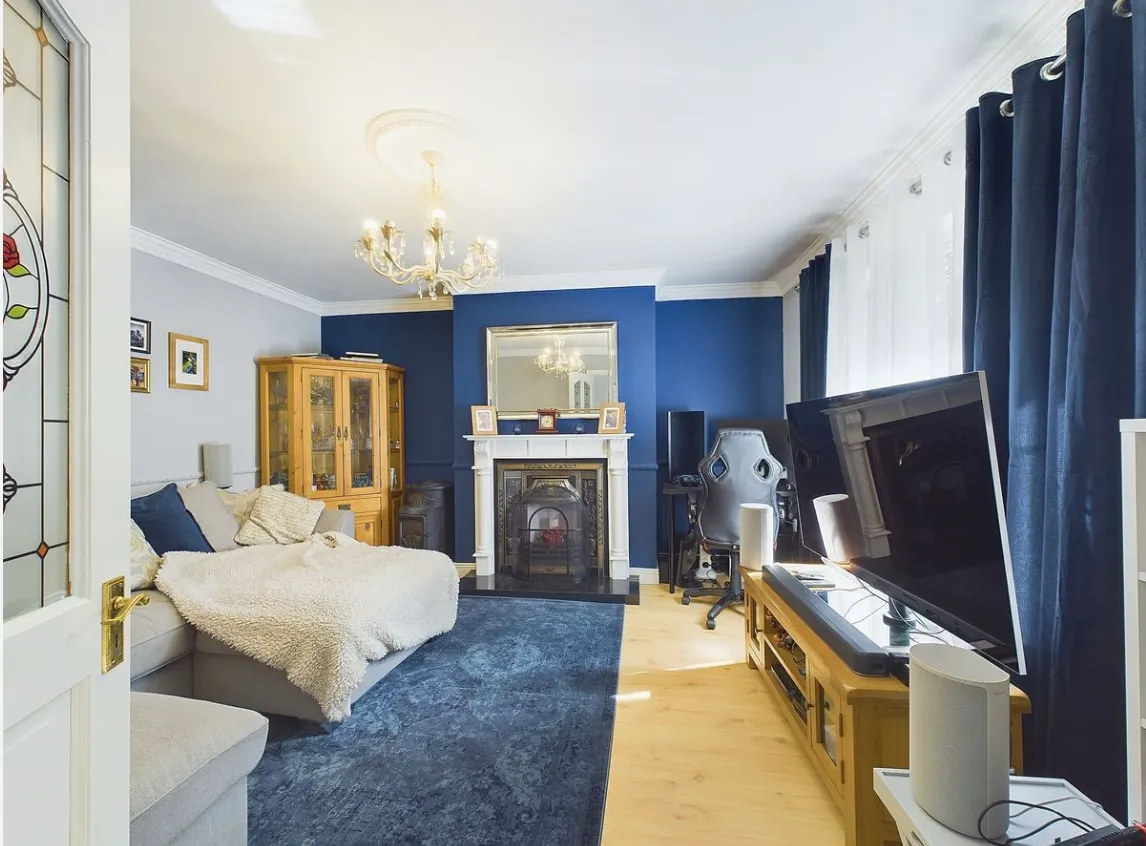 Living room with a sofa and throw blanket, fireplace with mirror above, large TV on a wooden stand, dark blue curtains and rug, wooden display cabinet and desk with an office/gaming chair.