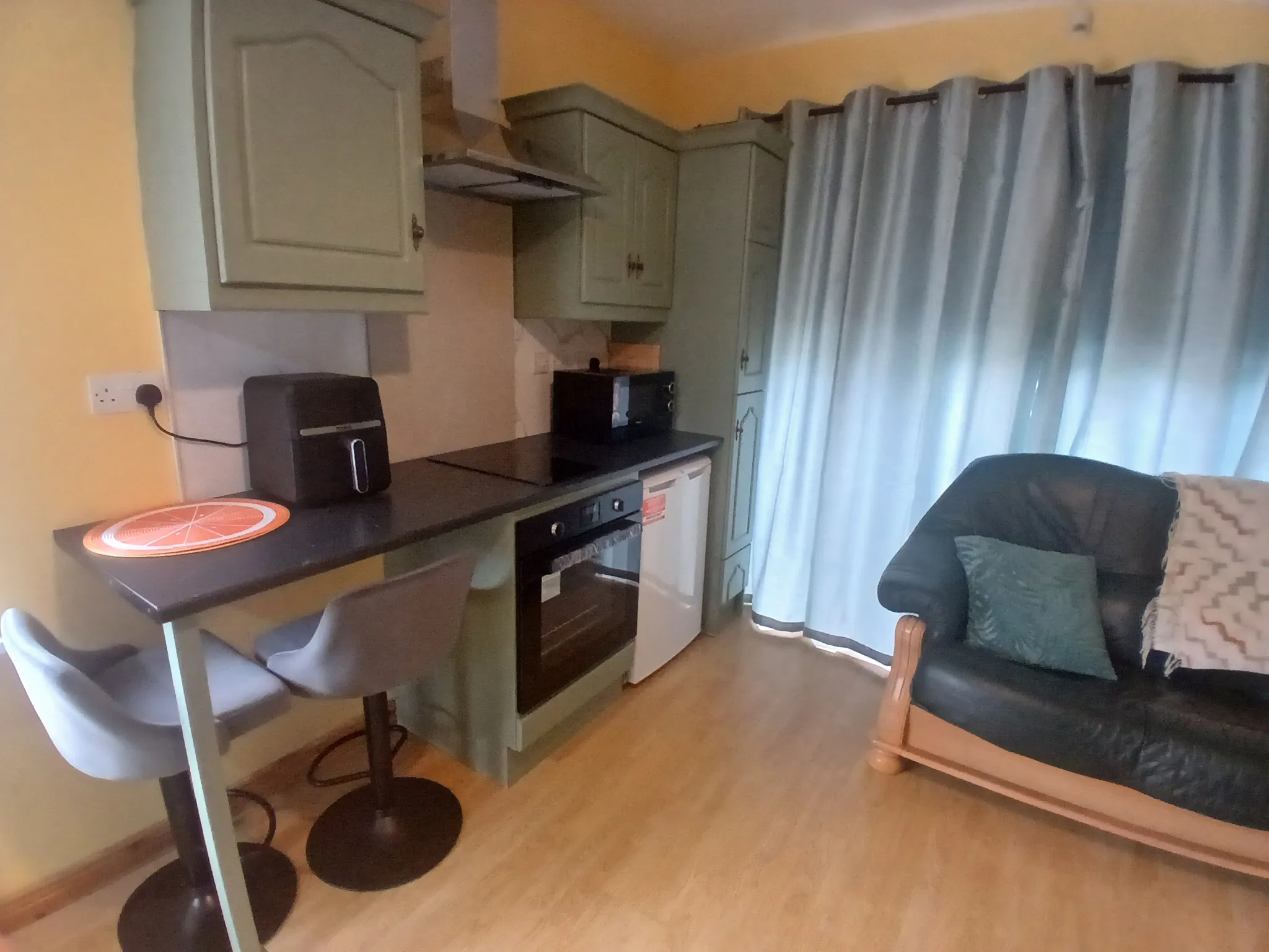 Kitchenette area with upper cabinets, countertop with toaster and microwave, built-in oven, two bar stools and curtains next to an armchair.