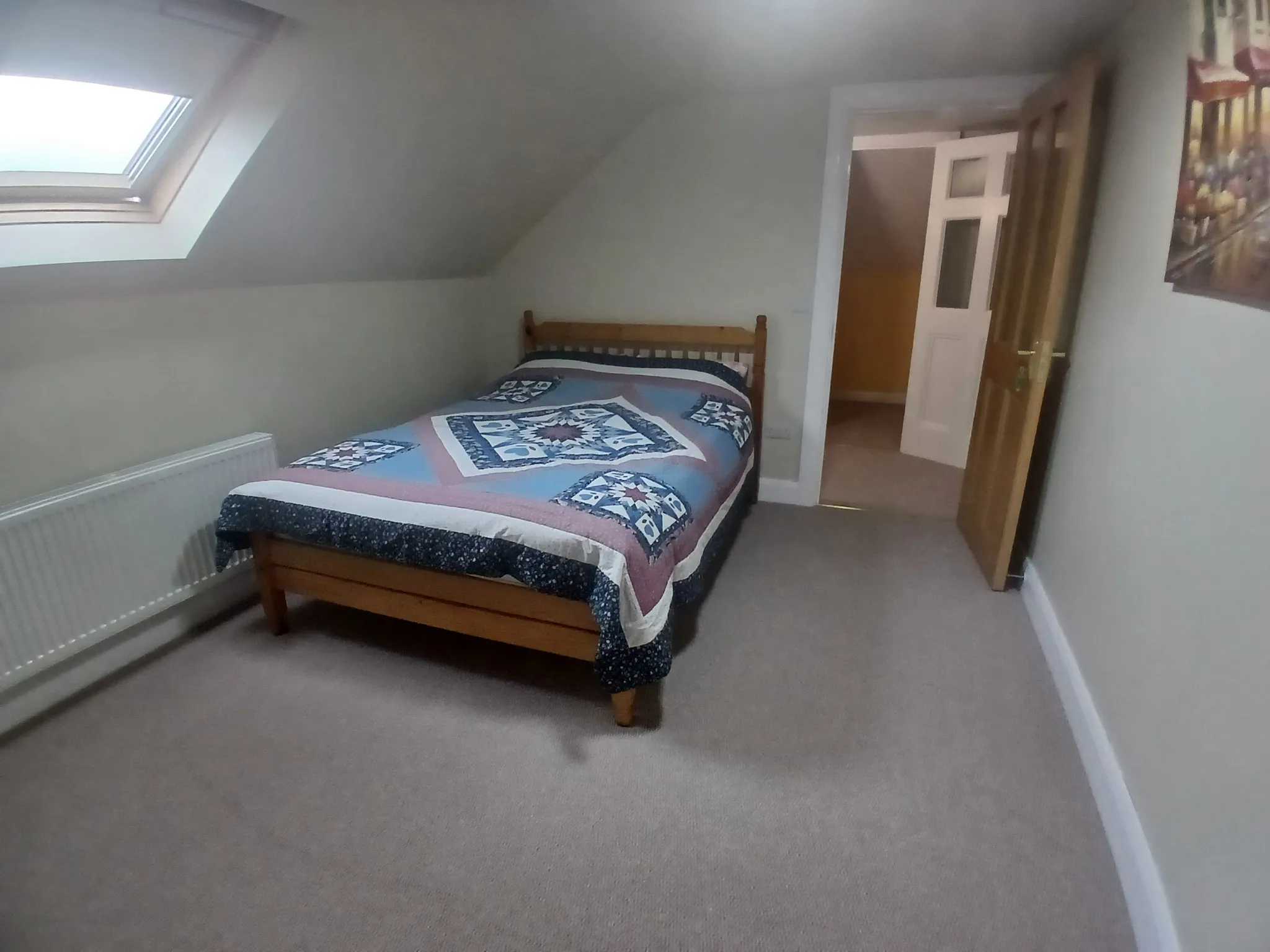 Small bedroom with a single wooden bed covered by a patterned quilt, skylight window in a sloped ceiling, radiator under the window and an open wooden door.