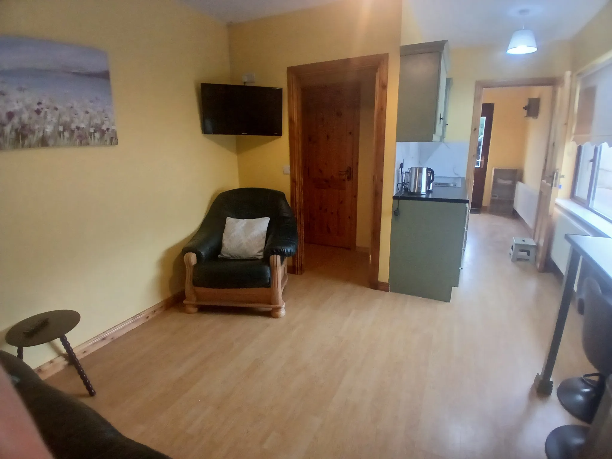 Living room area with an armchair, wall-mounted TV, wooden door and view into a small kitchen and hallway with windows.