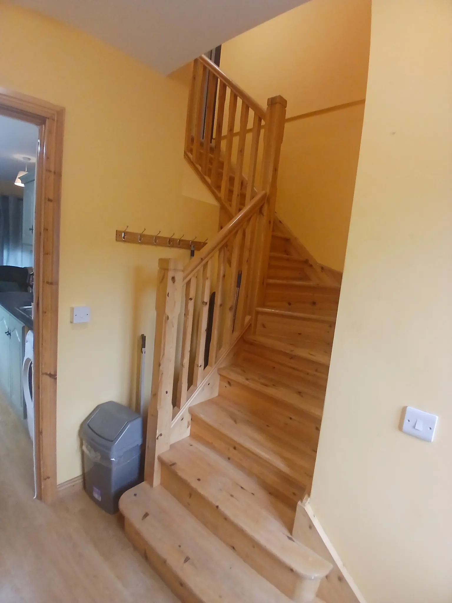 Hallway with a wooden staircase and handrail, coat hooks on the wall and a trash bin near a doorway.