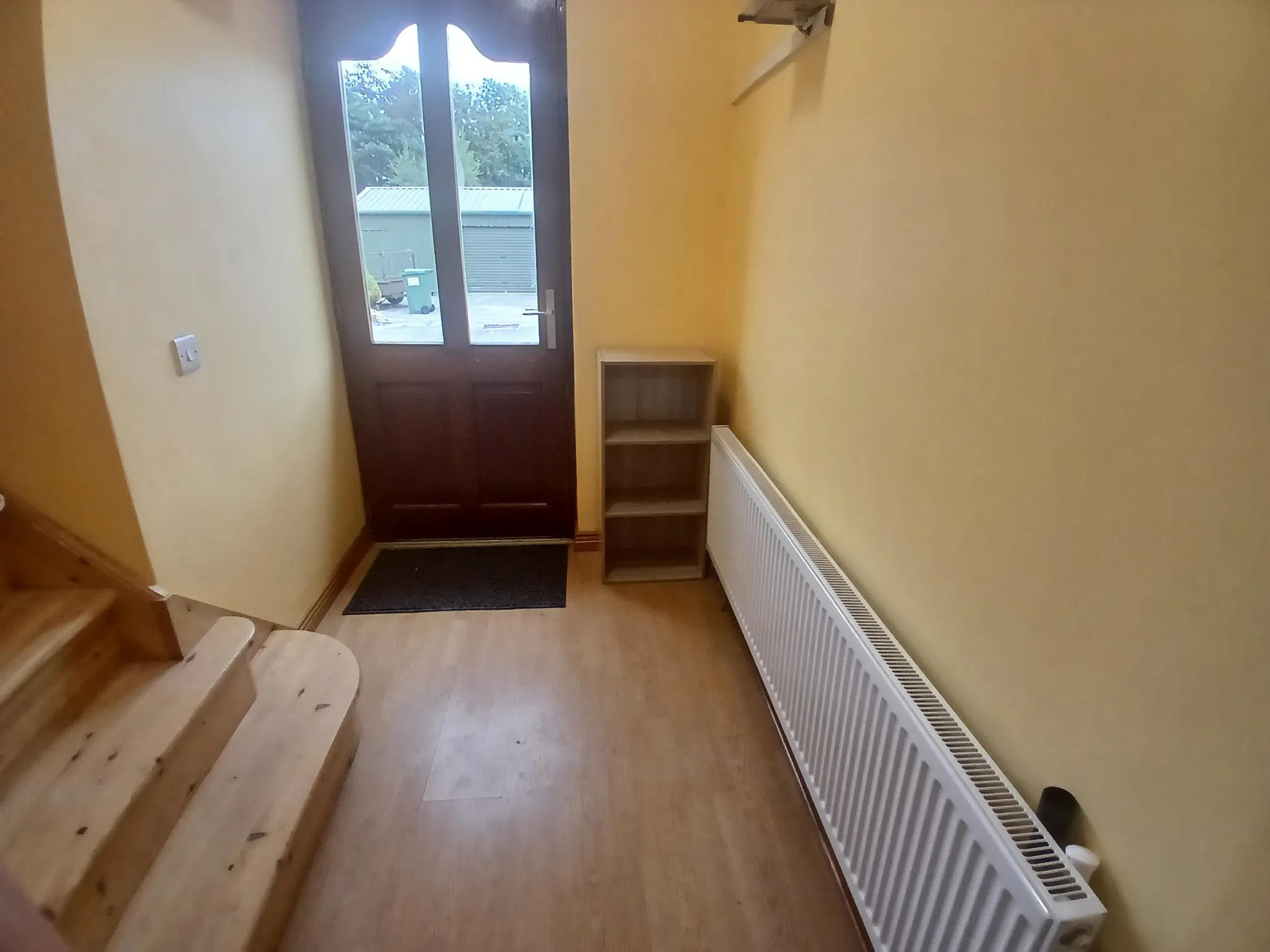 Narrow hallway with a wooden front door with glass panels, wooden stairs at left, a radiator along the right wall and a small open shelving unit beside the door.
