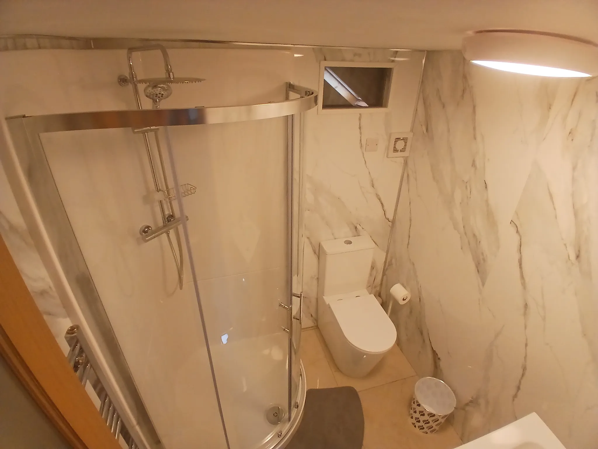 Bathroom with a corner glass shower enclosure, wall-mounted rain shower head, toilet, wall-mounted vent, and a small waste bin