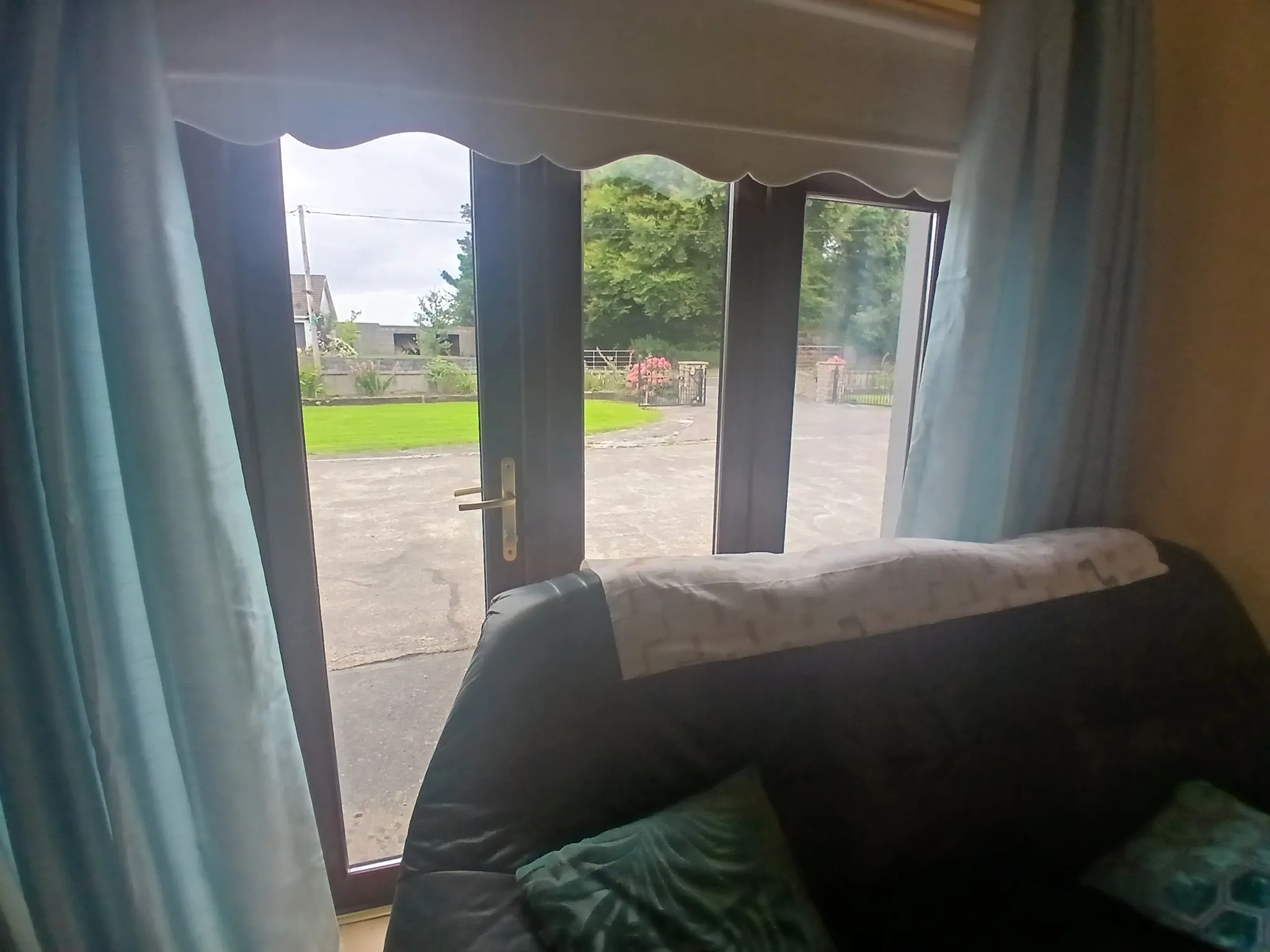 Living room with a sofa in front of glass patio doors and curtains, showing a view of a driveway and garden outside.