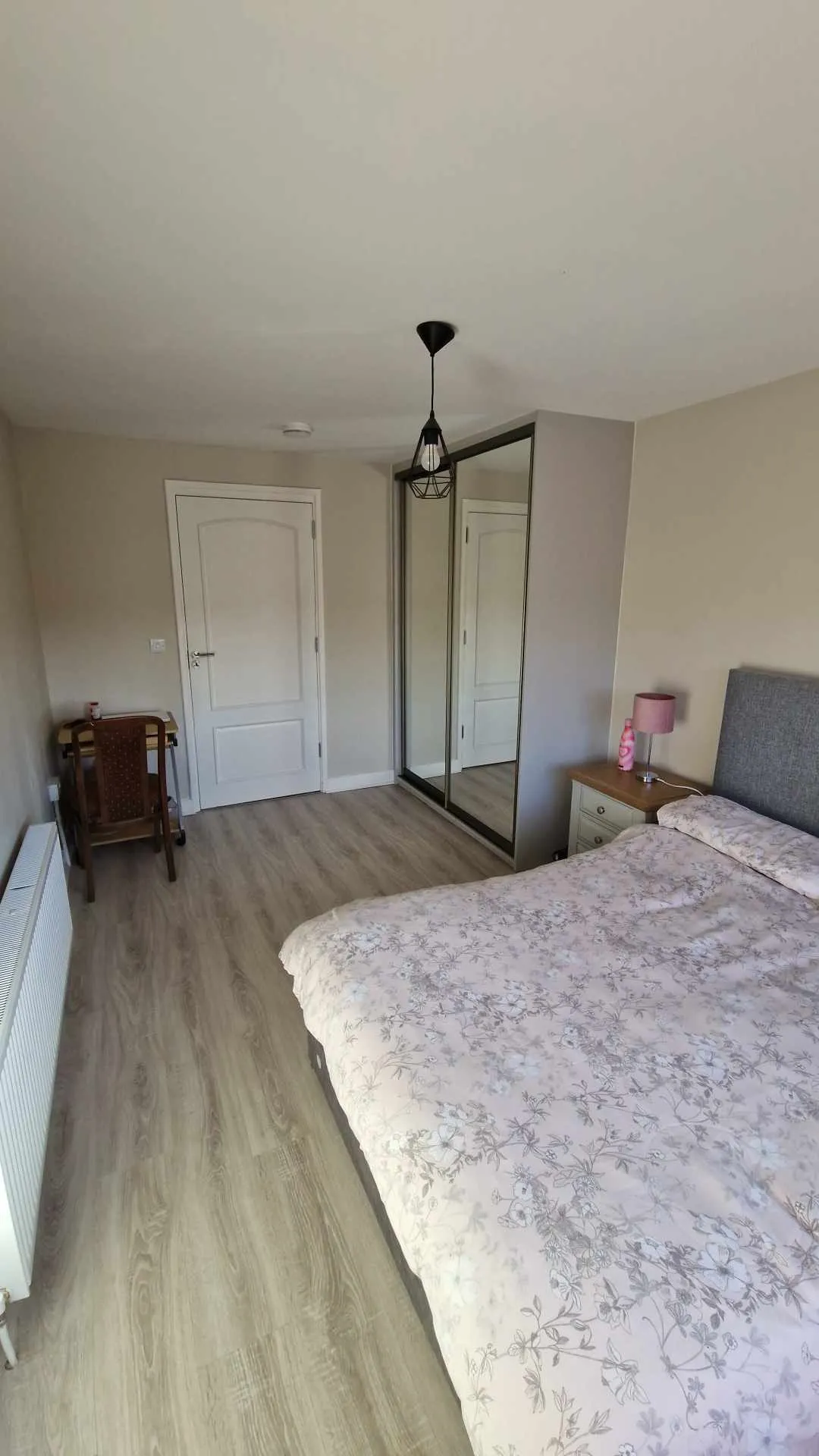 Bedroom with a double bed, bedside table and lamp, mirrored sliding wardrobe, small desk and chair, pendant ceiling light and radiator.