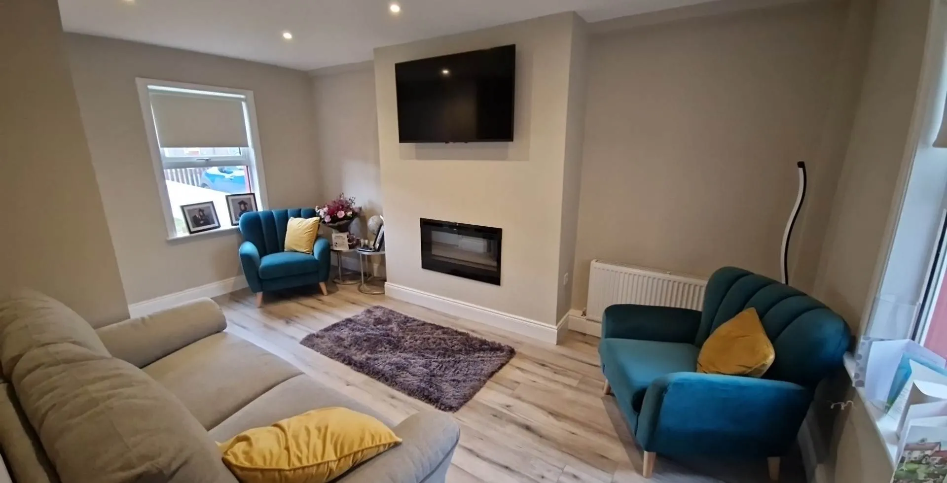 Living room with a wall-mounted TV and fireplace, two teal armchairs with yellow cushions, a beige sofa, small rug, and window.