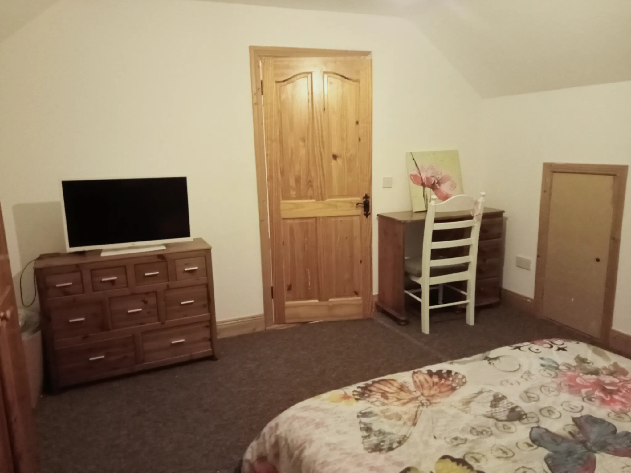 Bedroom corner with a wooden door, dresser with a TV, a desk and chair, and a bed partially visible in the foreground.