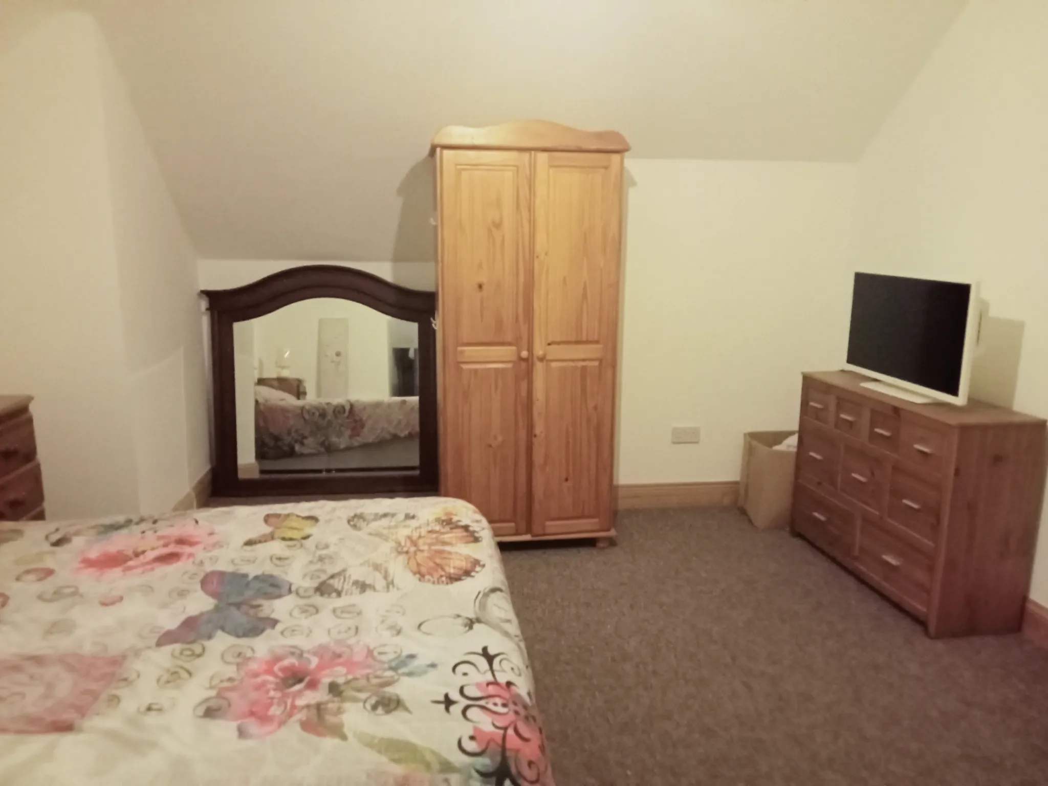 Bedroom with a floral bedspread, wooden wardrobe, large framed mirror and dresser with a TV on top
