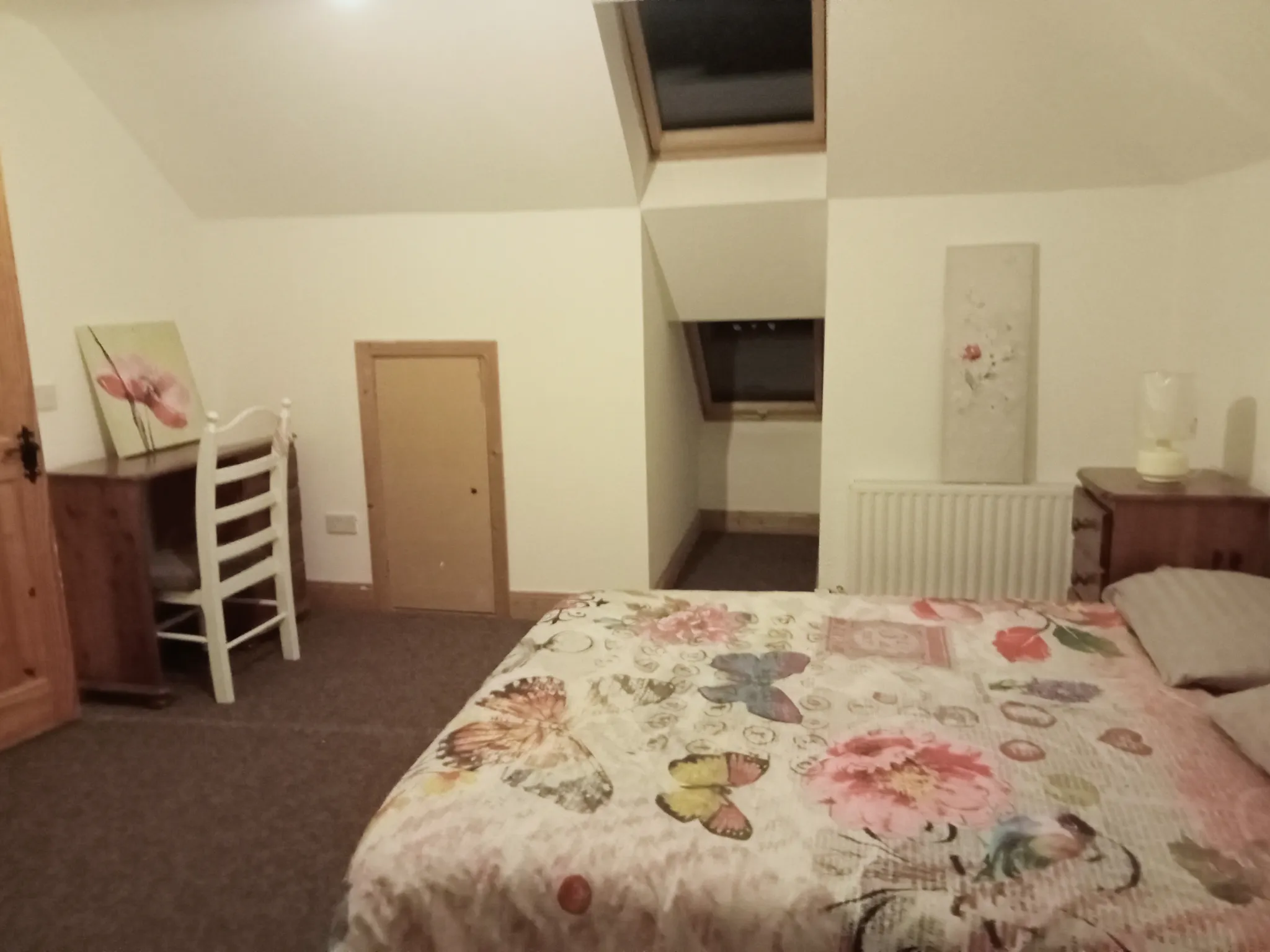 Bedroom with a floral bedspread, visible bedside table and lamp, a chair and desk, small attic-style window in a sloped ceiling and a radiator.