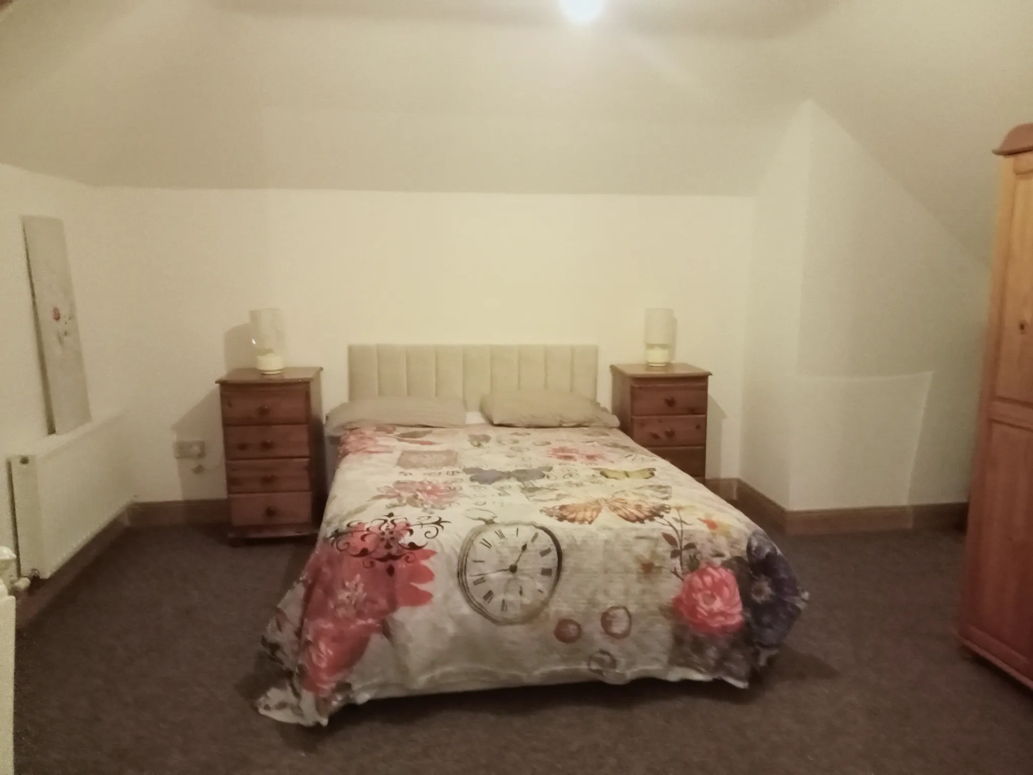 Bedroom with a double bed, floral duvet, padded headboard, two wooden bedside tables with lamps, a wardrobe on the right and a radiator on the left.