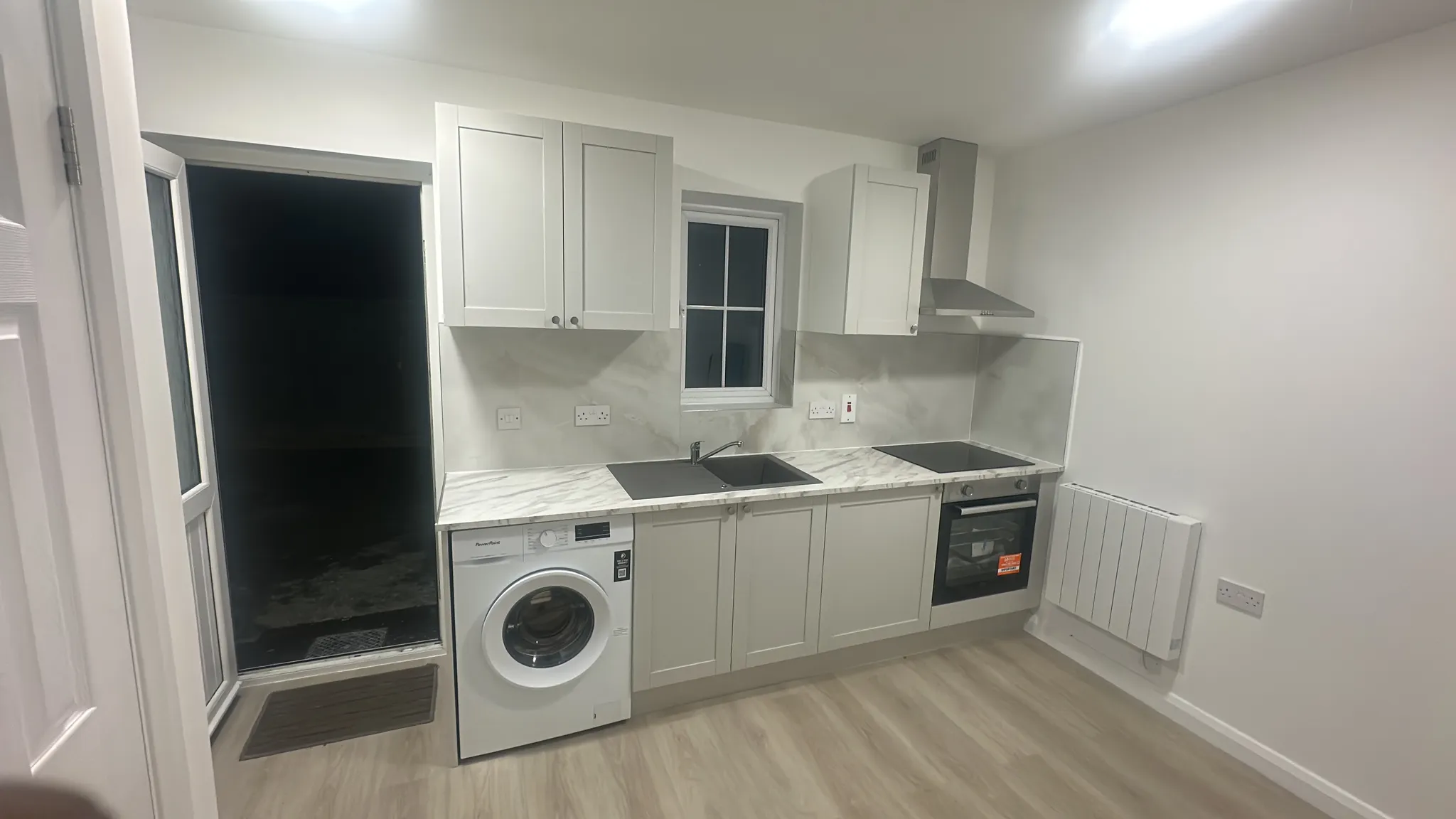 Kitchen with white cabinetry, marble countertop, black induction hob, built-in oven, washing machine, and window with dark exterior view