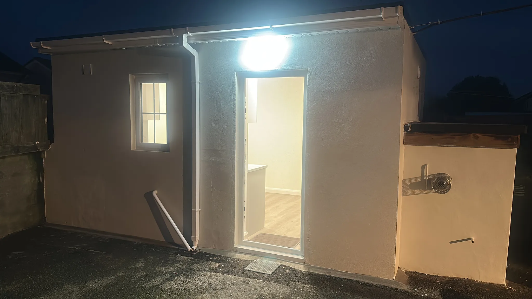 Exterior of a small modular residential unit at night, illuminated by overhead lighting, with white walls, a glass door entrance, and a window visible on the left side.