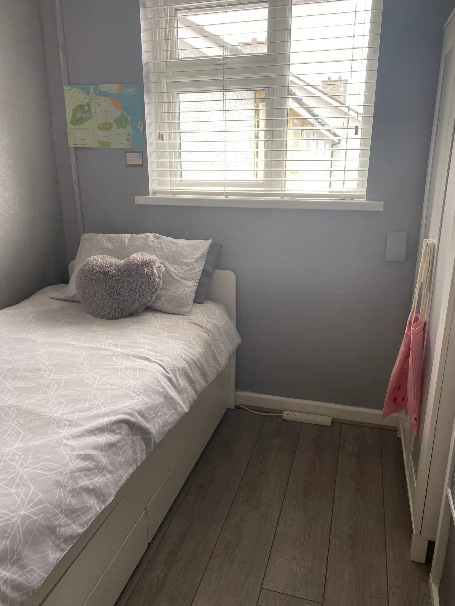 Small bedroom with a single bed against a wall, heart-shaped cushion on the bed, window with horizontal blinds and light grey walls.