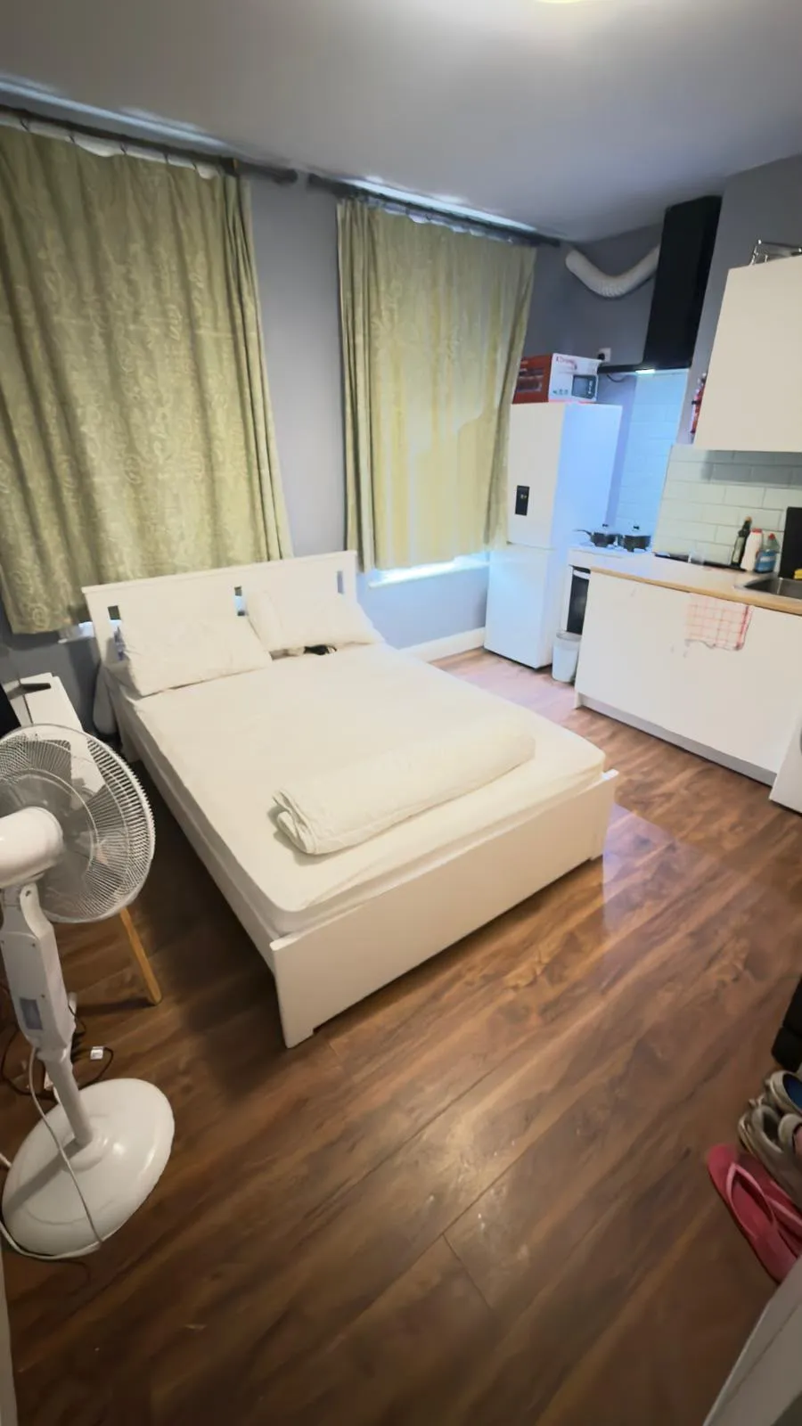 Studio bedroom with a double bed, two pillows, a standing fan, curtains over a window, and a small kitchenette with refrigerator, sink and cabinets