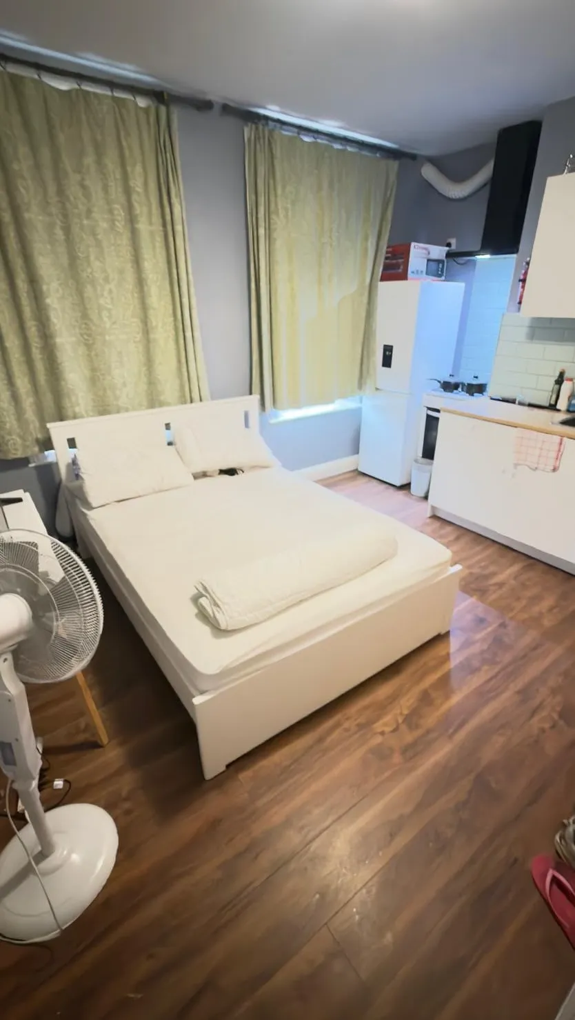 Bedroom with a double bed, pillows and folded duvet, a standing fan, curtained windows and a small kitchenette with fridge and stove visible