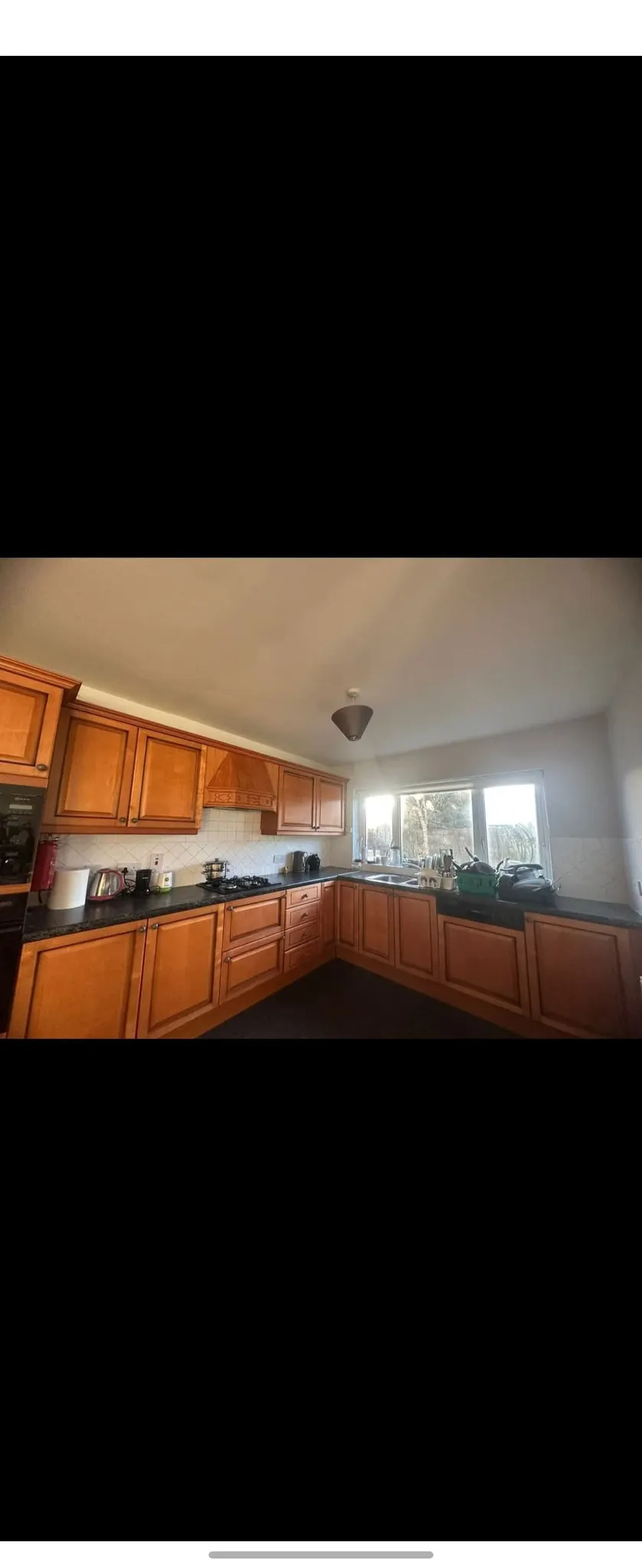 Kitchen with wooden cabinets and black countertop in an L-shape, gas stovetop, sink beneath a window and overhead light fixture.