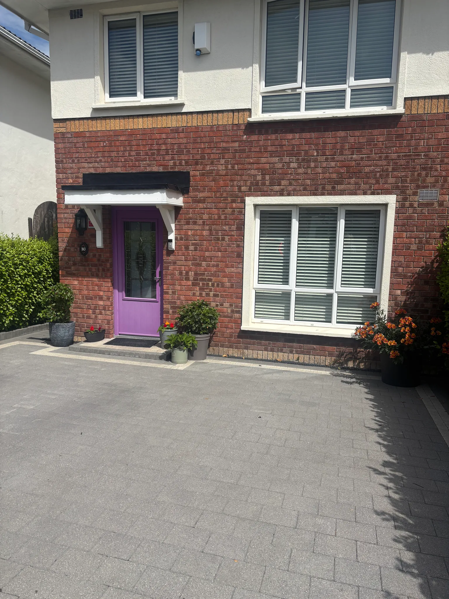 Exterior view of a two-story brick house front with a purple entrance door, white-framed windows with blinds, small porch awning and potted plants by the doorway.